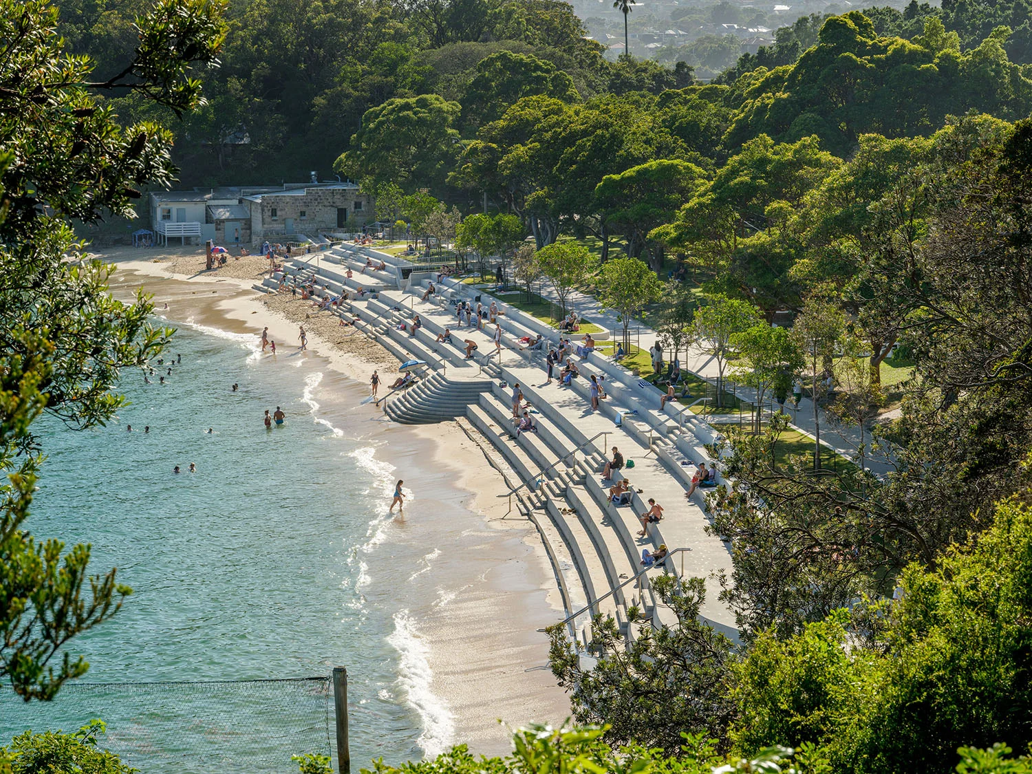 A photograph taken looking down the length of a beach seawall on a sunny day. The image is framed by green trees.