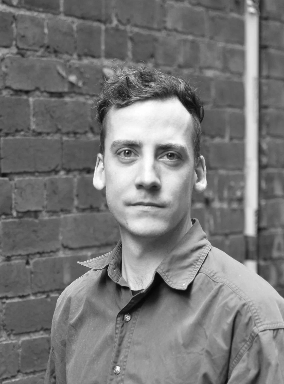 A black and white headshot of a person standing in front of a gritty, dark brick wall.