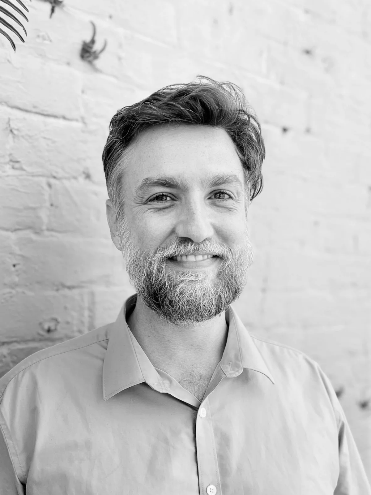 A black and white headshot of a smiling person with a beard in front of a white brick wall.