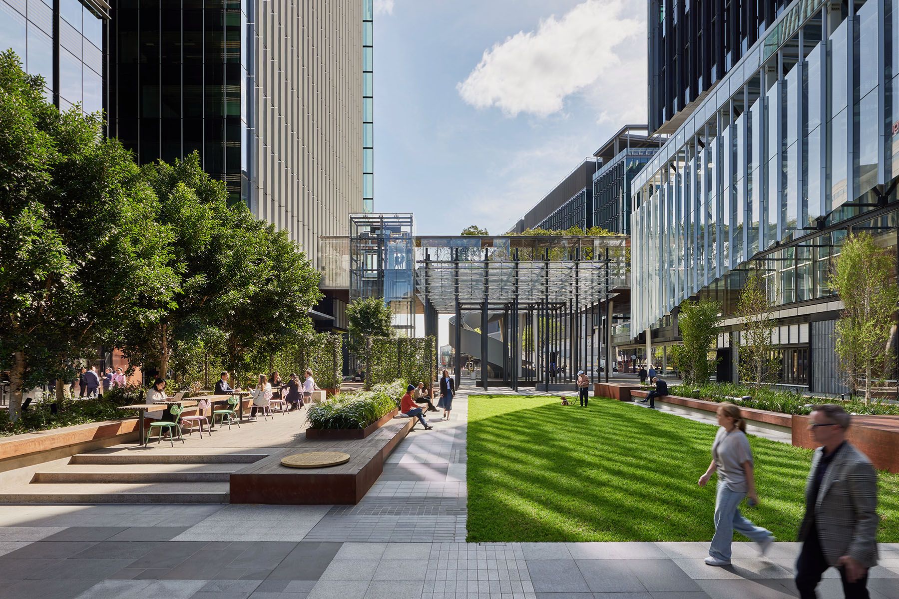 A photo of an urban plaza, surrounded by buildings, with trees, seating, a lawn, and a pavilion and elevated park in the background. 