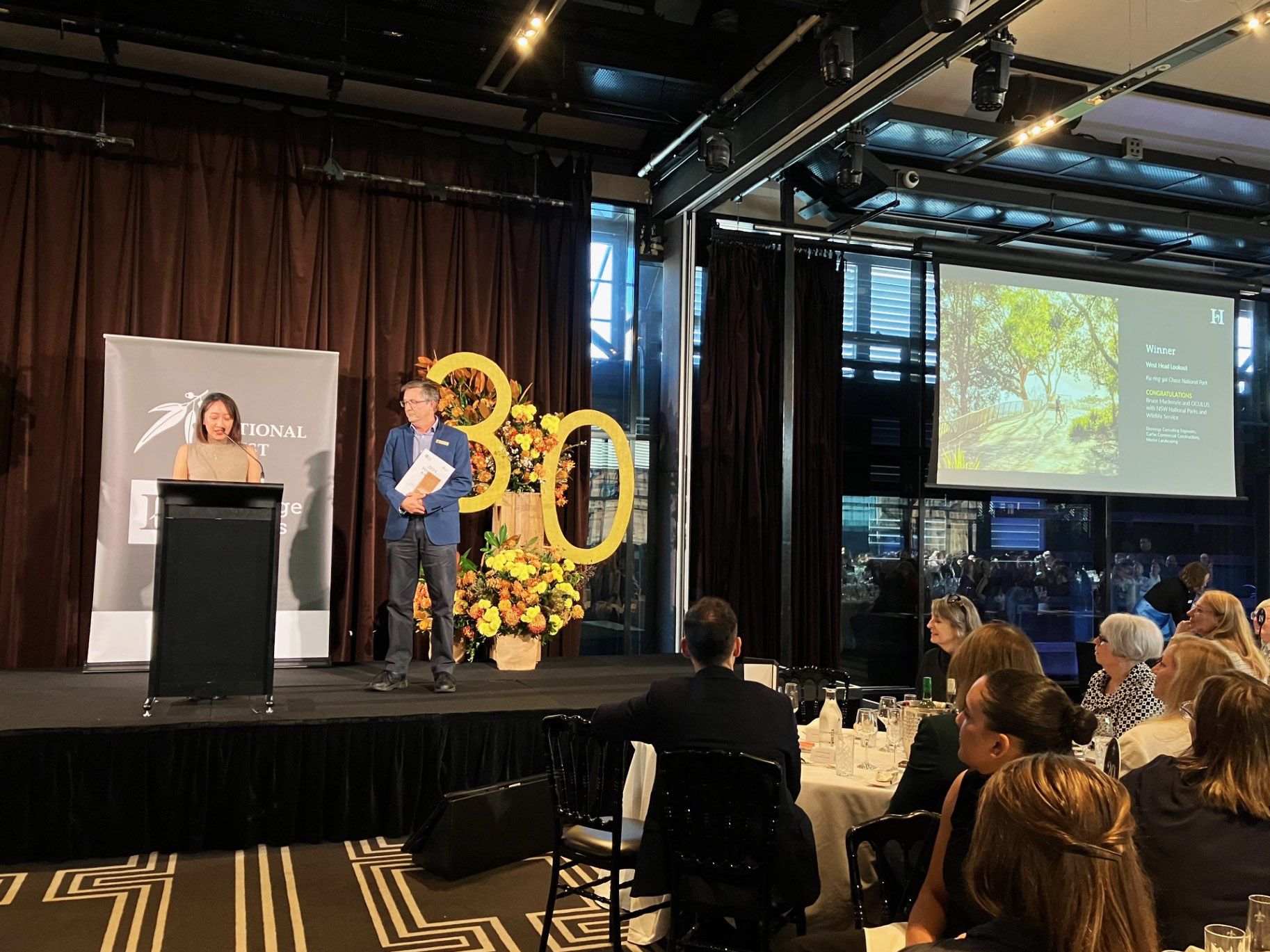 A photograph of a seated awards ceremony, two people are on stage collecting a trophy. There is a floral arrangement with a big "3" "0" cut out. 