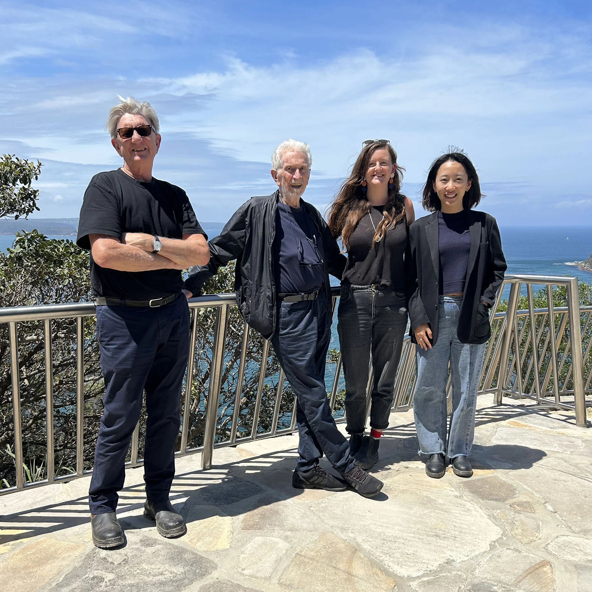 A photo of four people standing on a sandstone lookout with blue water and skies behind. They are all smiling, and wearing black and shades of blue.
