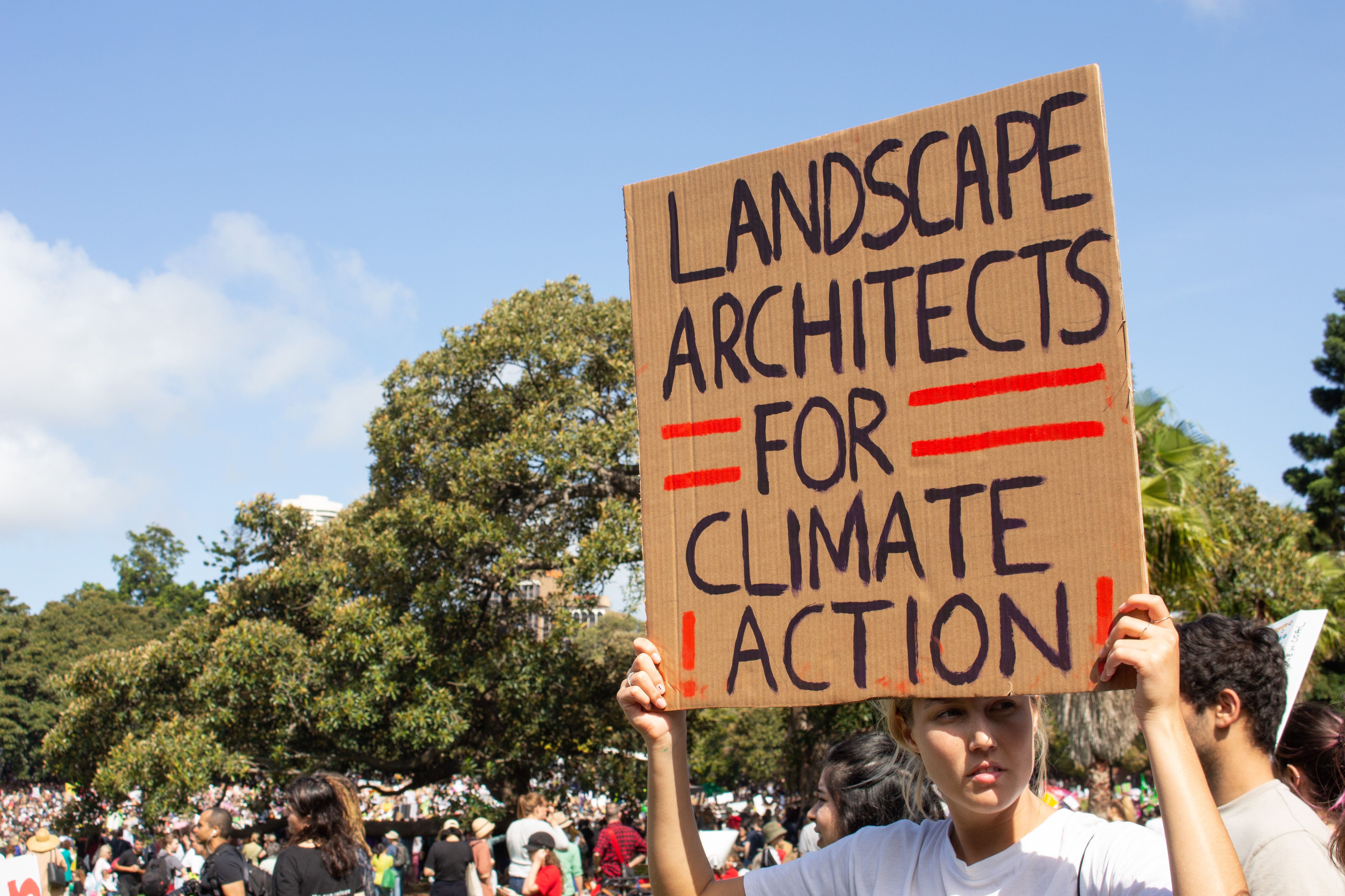 A photograph of a landscape architect at a climate rally, holding a cardboard sign above their head that says 'Landscape Architects for Climate Action'