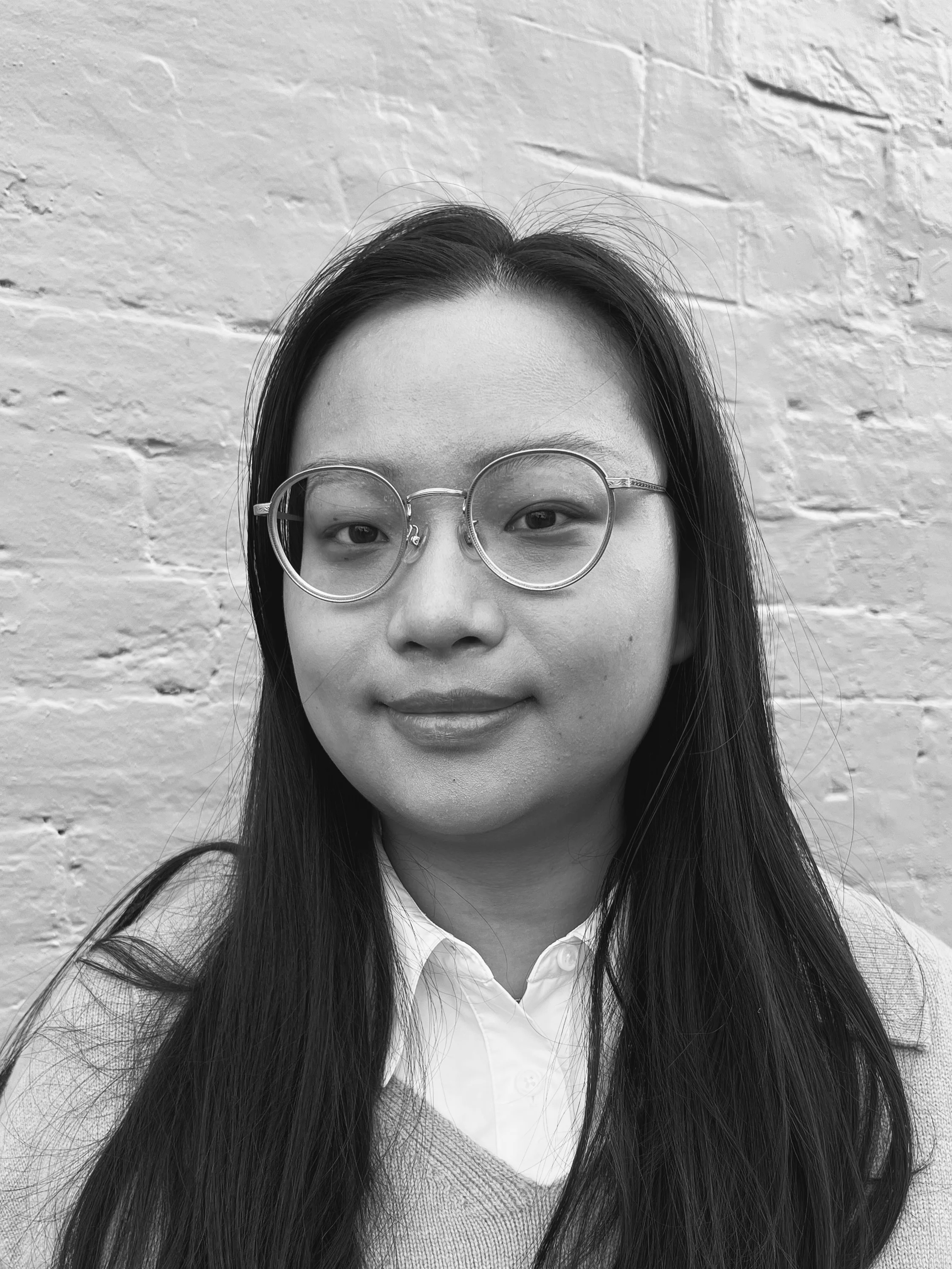 A black and white headshot of a person with long hair. They are wearing a white shirt and jumper, and standing in front of a painted brick wall.