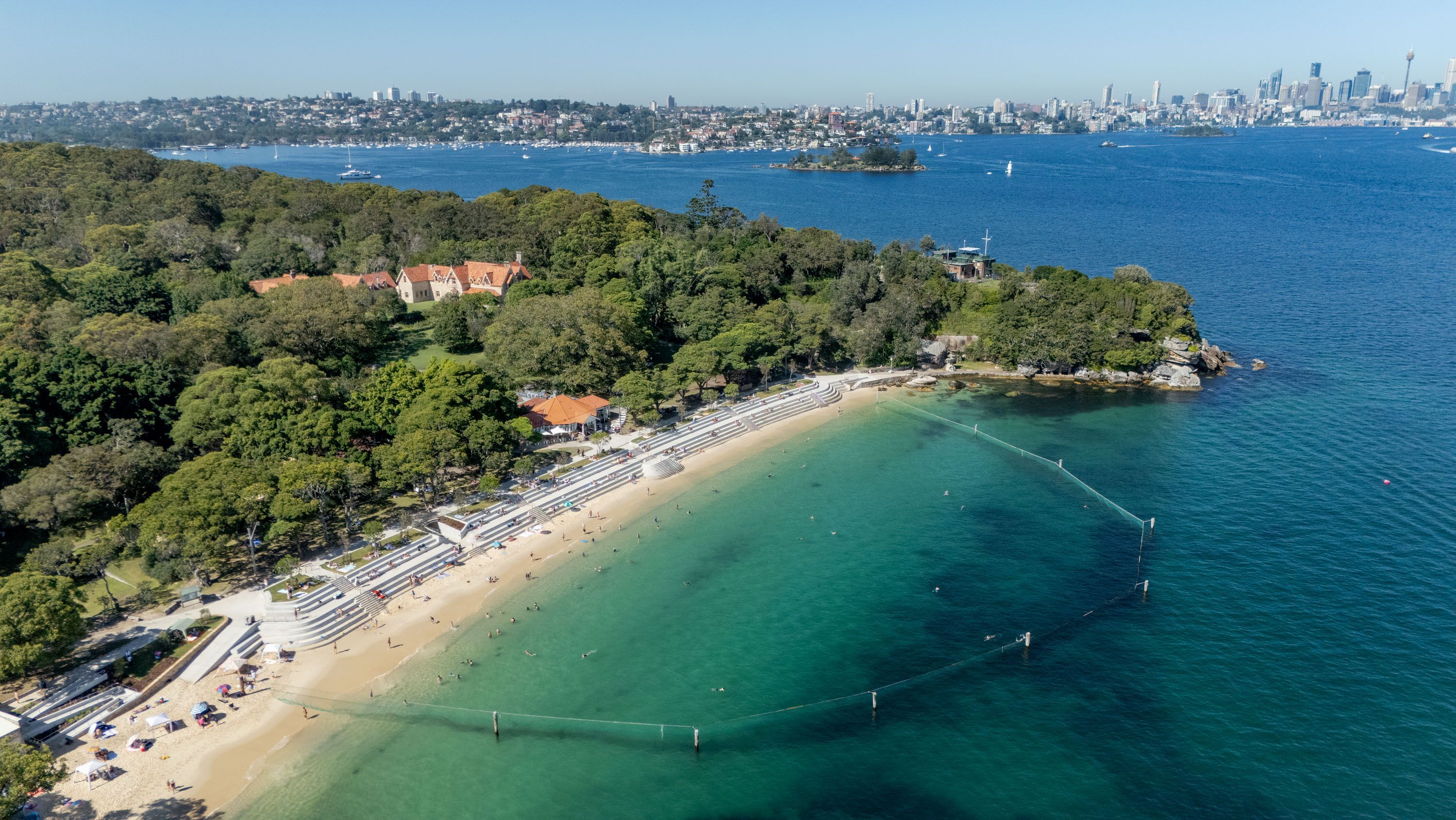 A drone photograph of a harbour beach with a shark net and new seawall, taken on a sunny day, with a city skyline in the distance,