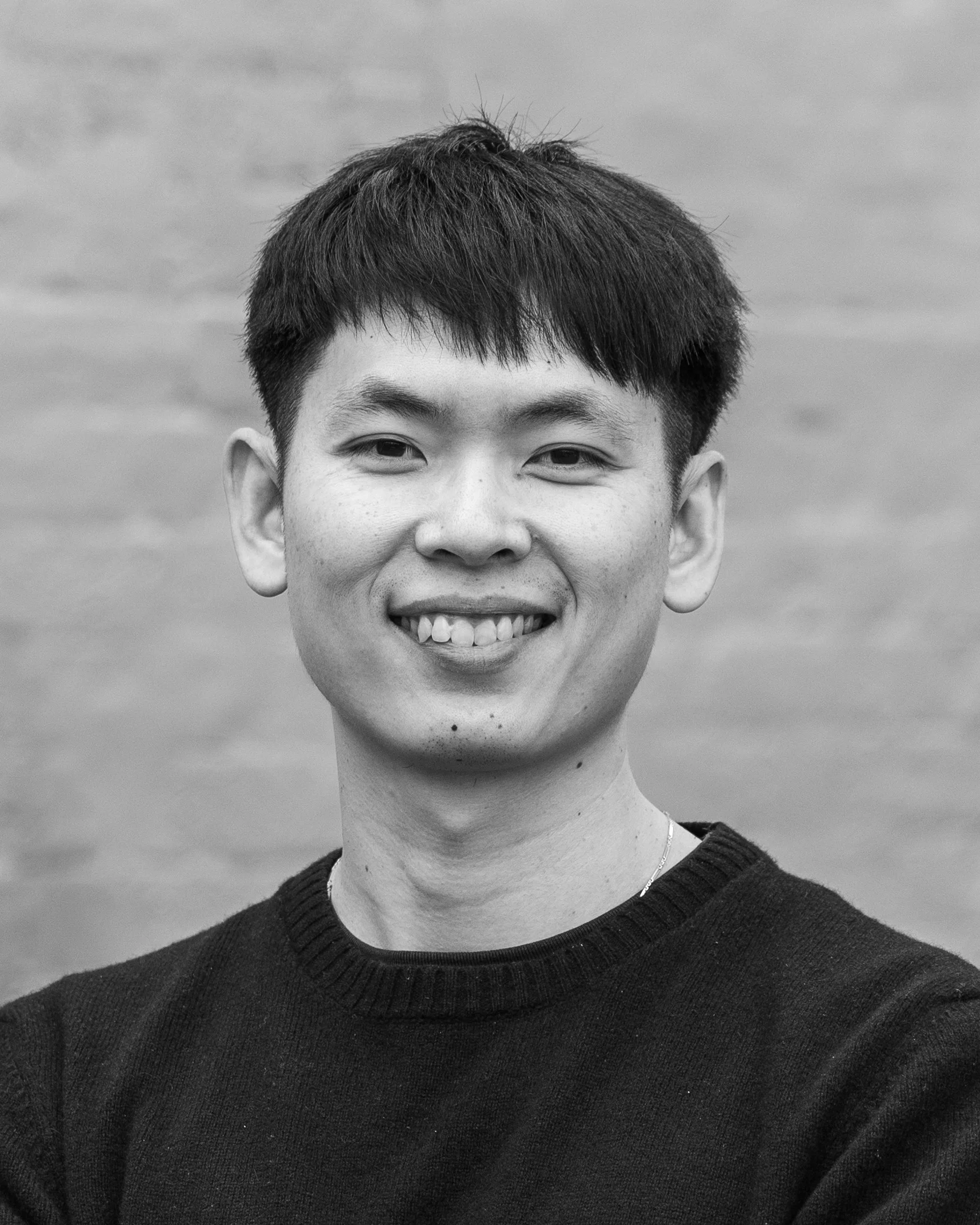 A black and white headshot of a smiling person in front of a brick wall.