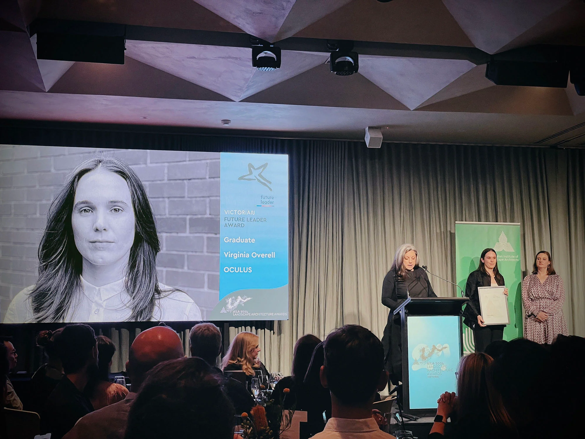 A photo of an awards presentation stage. There is a black and white headshot on a projector screen, and three people on stage - one is speaking at a lectern, and one is holding a framed certificate.