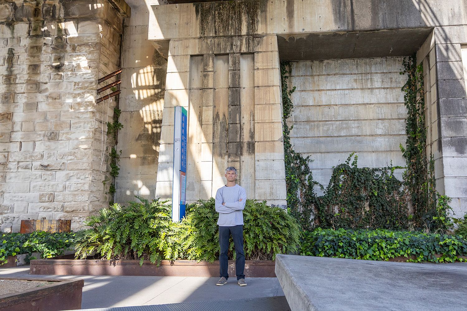 Man in casual clothes stands with crossed arms with vegetation and bridge infrastructure behind him.