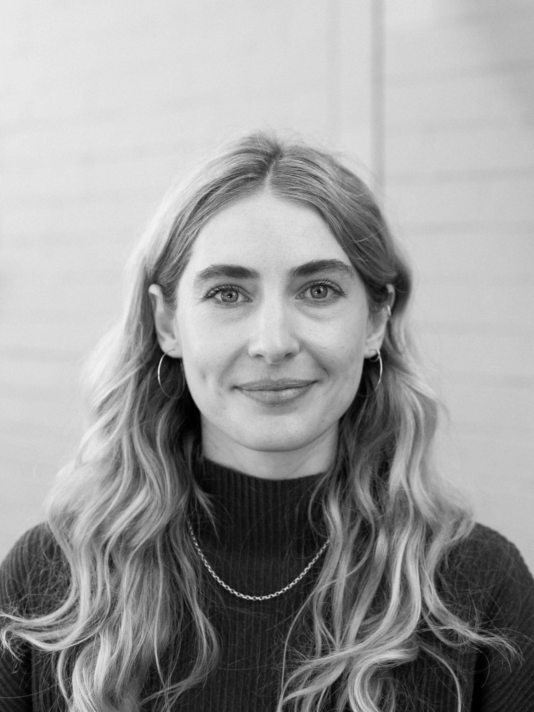 A black and white headshot of a person with long hair in front of a brick wall pained white.