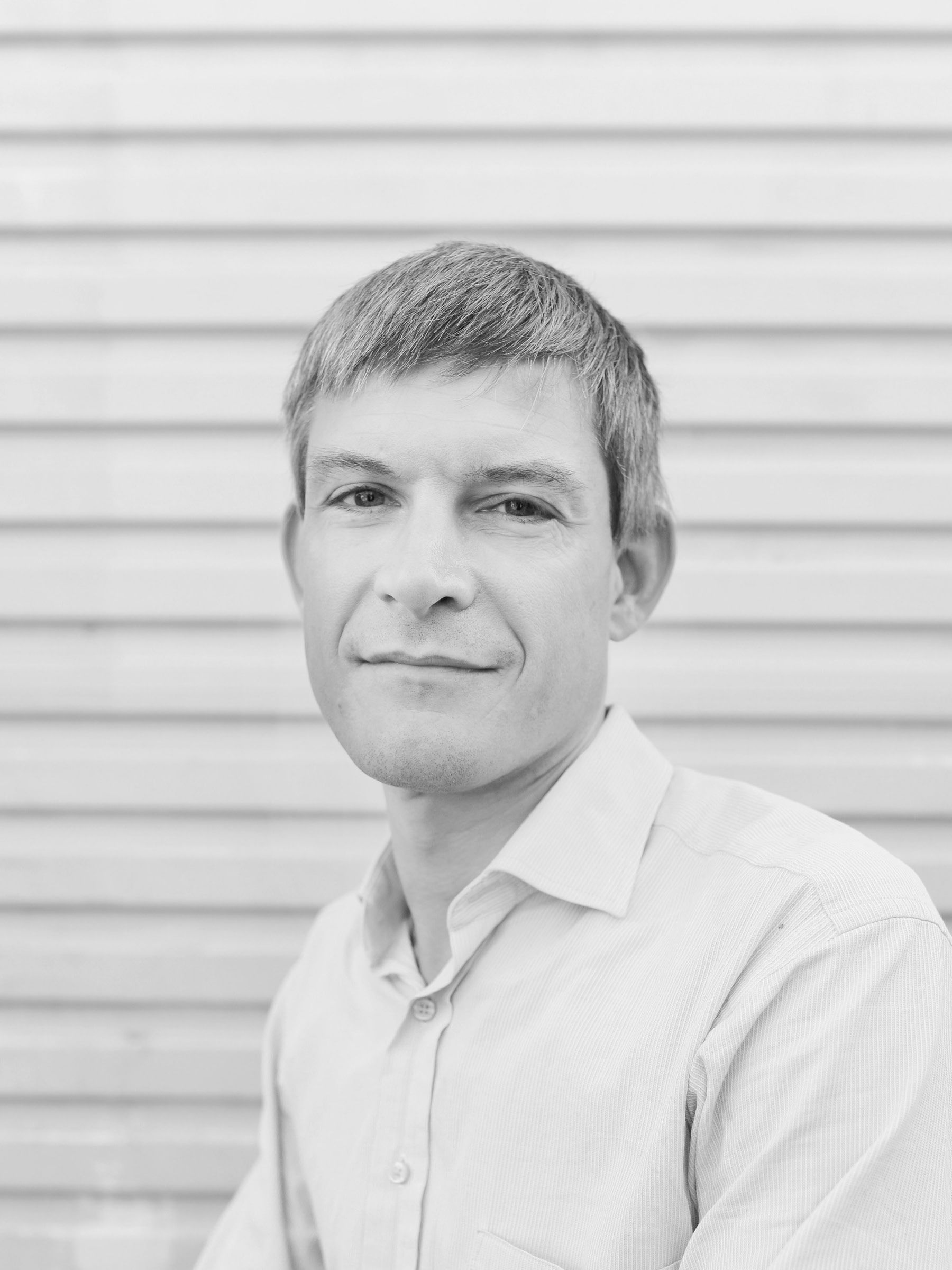 A black and white headshot of a person looking at camera, wearing a white shirt, in front of a metal roller door.
