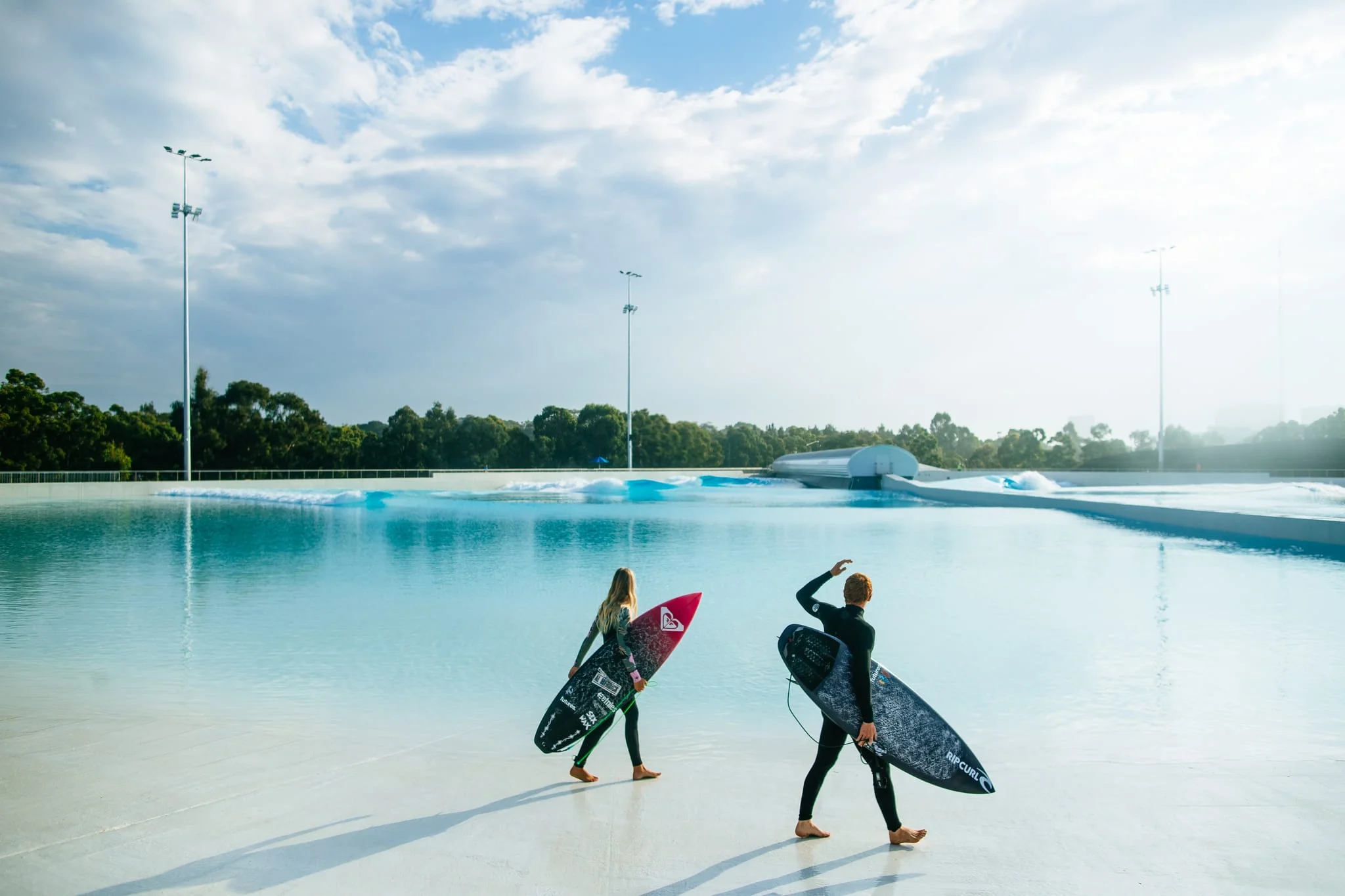 A photograph of two people carrying surfboards while walking on the shore of a large wave pool.