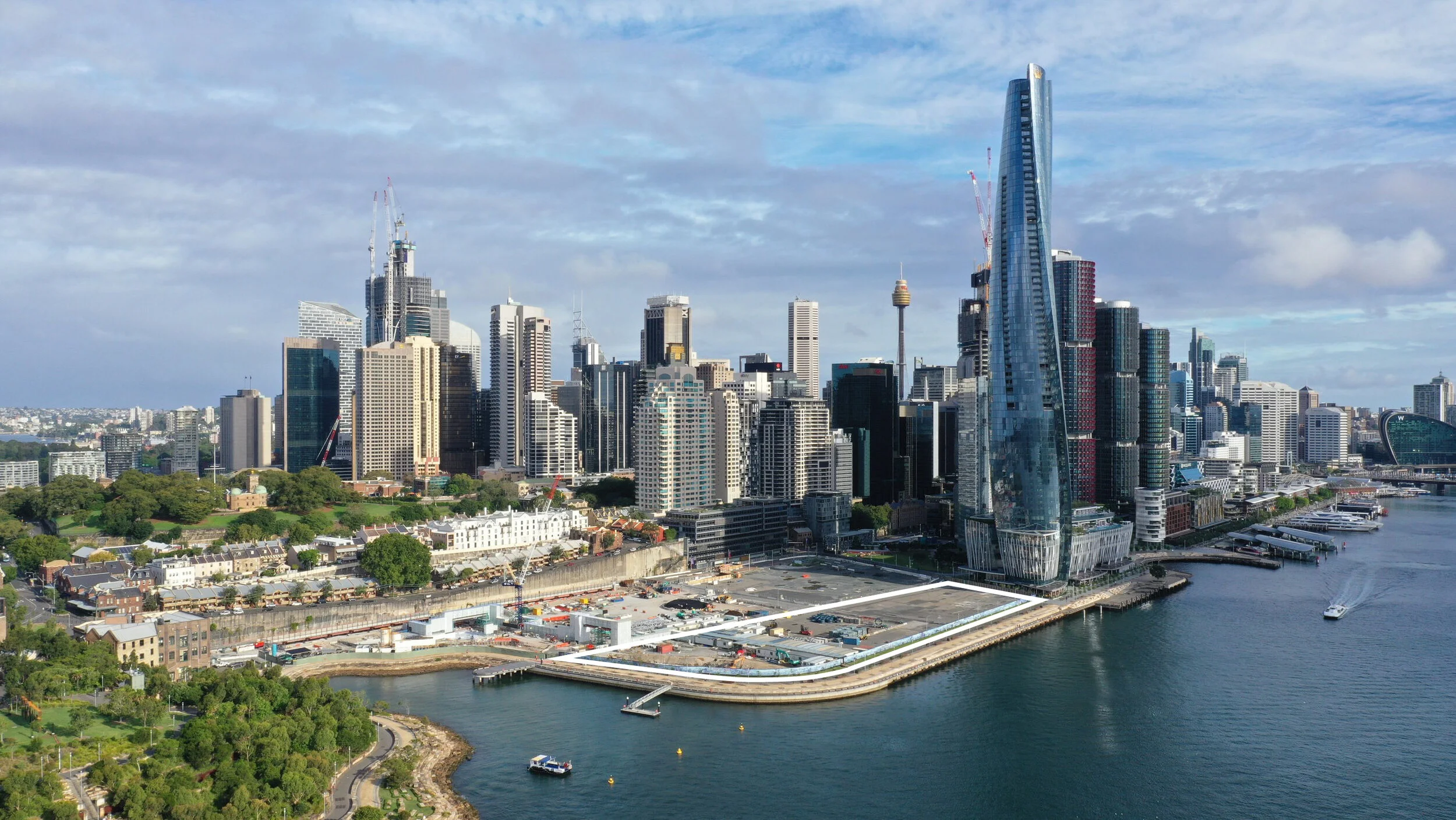 An aerial photograph of Sydney, with the harbour and Barangaroo Precinct in the foreground, and the city and a cloudy sky behind. A white outline has been added to indicate the Harbour Park site perimeter, as part of a larger construction site.