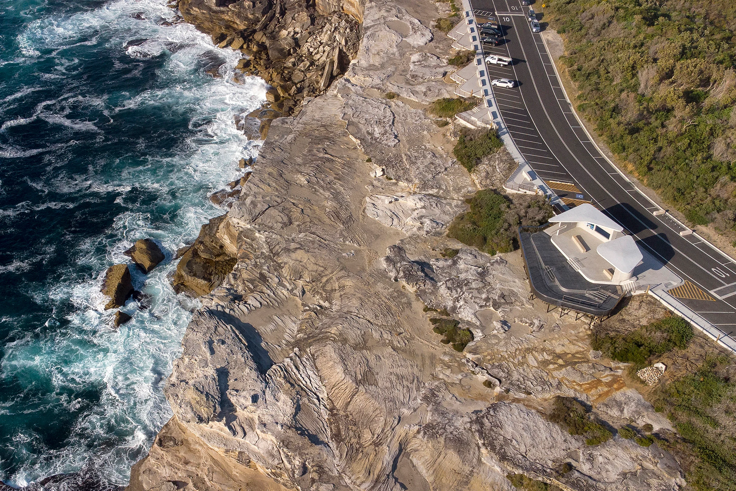 An aerial photograph of a rocky coastline. On the left of the image, waves crash against cliffs. On the right of the image a viewing platform and road is visible.