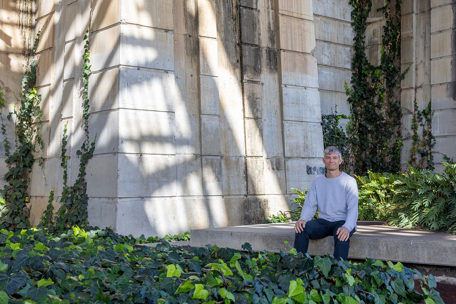 Man in casual clothes sits on concrete base with bridge infrastructure behind him.