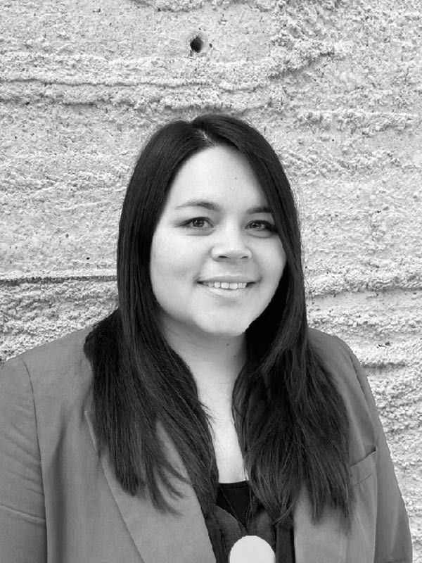 A black and white headshot of a smiling person in front of a heavily textured white wall. 