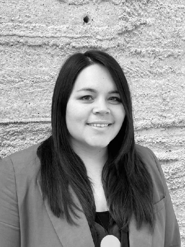 A black and white headshot of a smiling person in front of a heavily textured white wall.