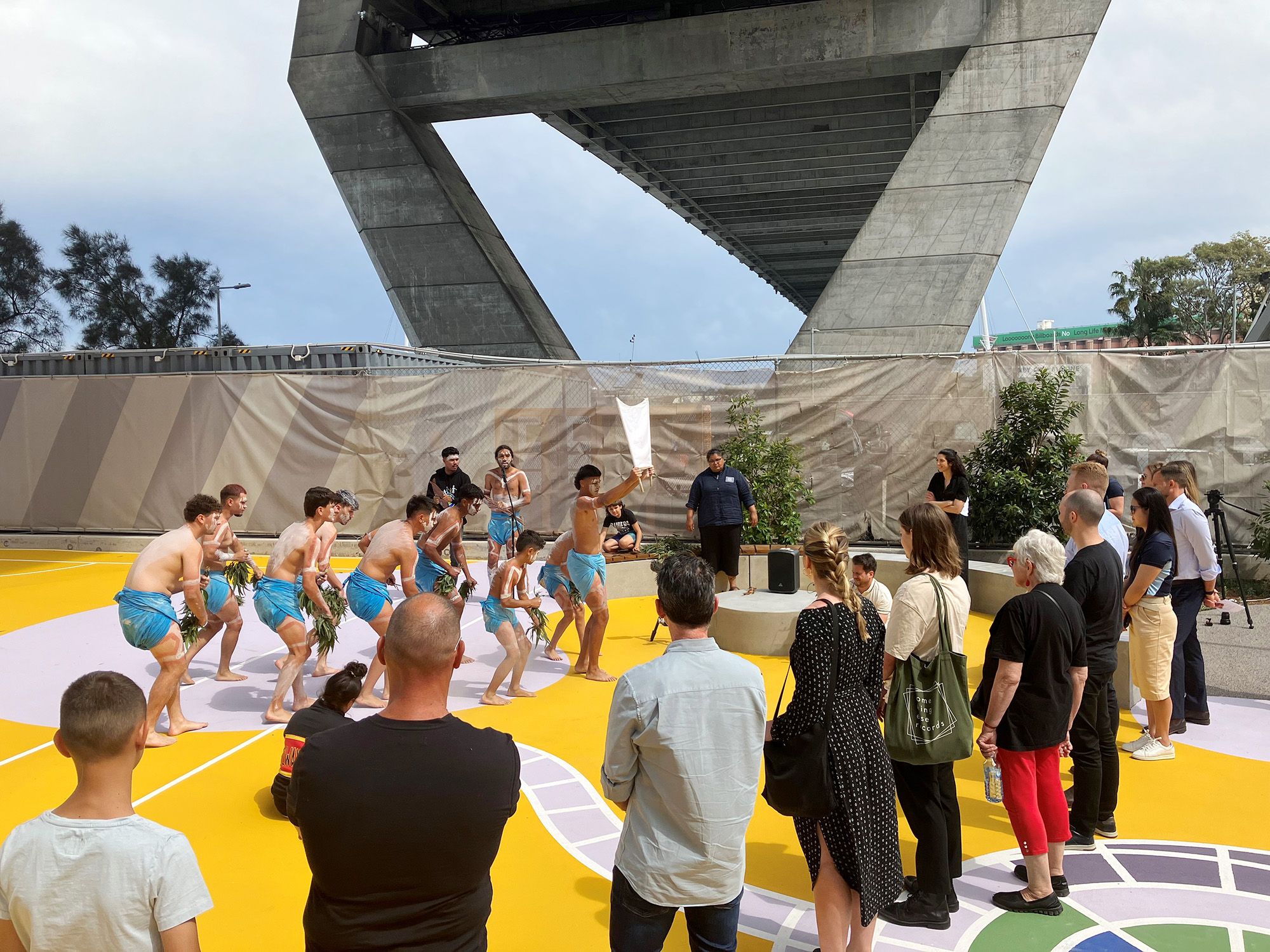 A photograph of a small crowd watching a group of dancers perform in a bright, urban, pop-up park. A large concrete bridge stands overhead. 