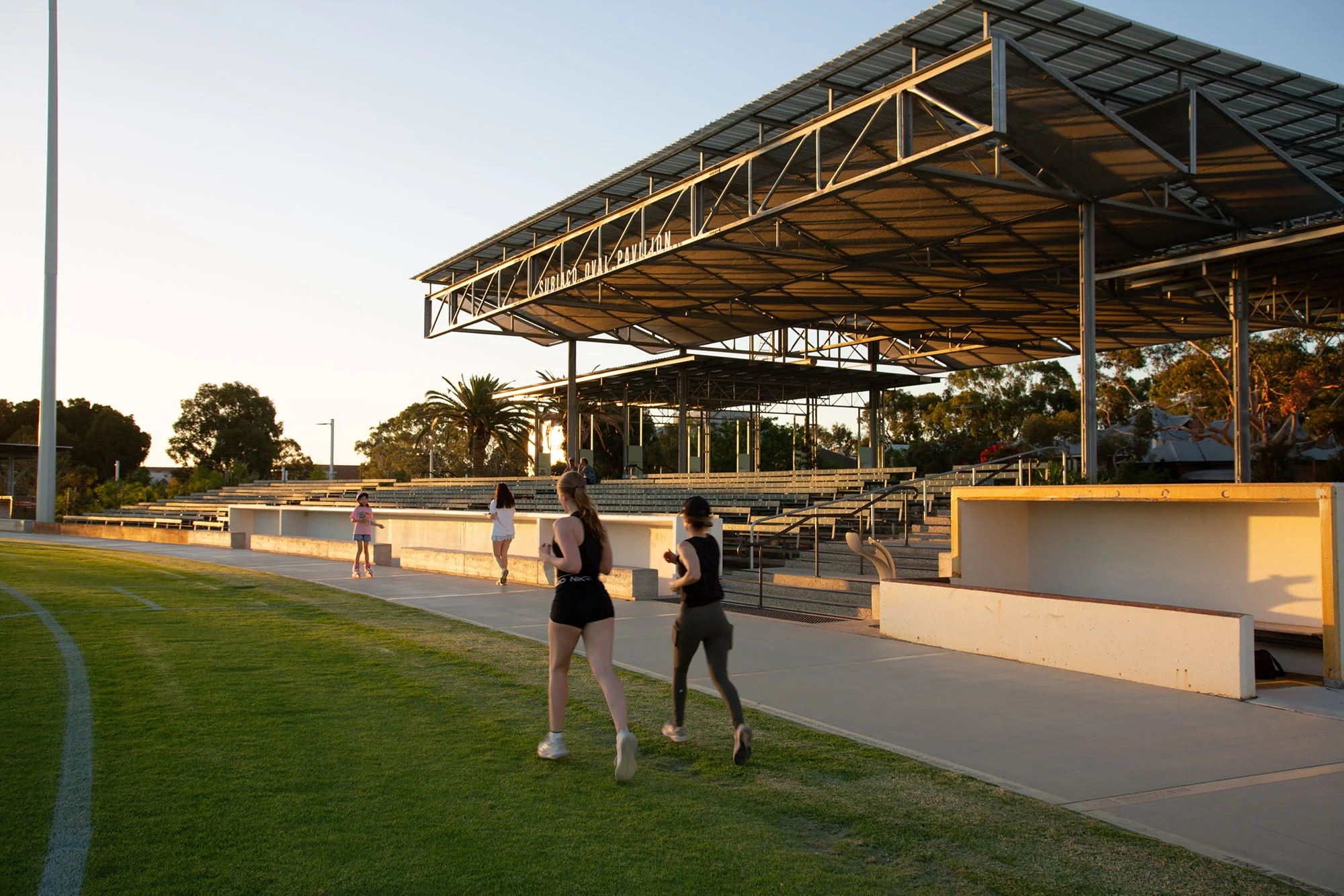 A photograph of people running around an oval at sunset, past a seating structure with a sign that reads "Subiaco Oval Pavilion"