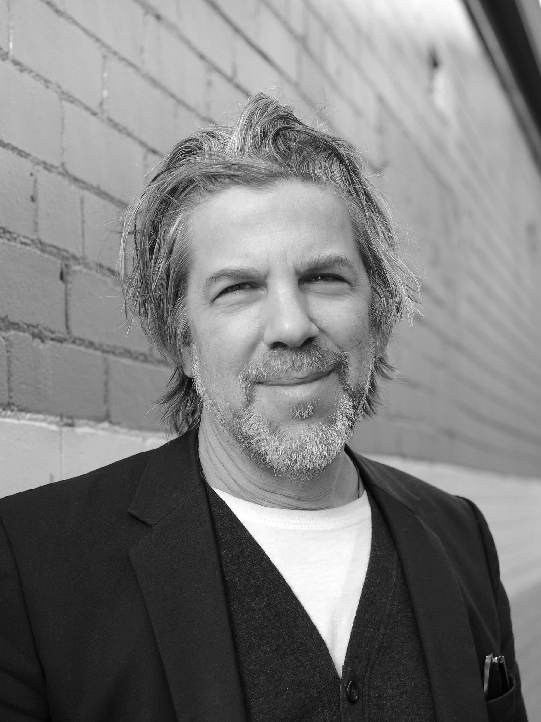 A black and white headshot of a person in a laneway. They have a pen in their top jacket pocket.