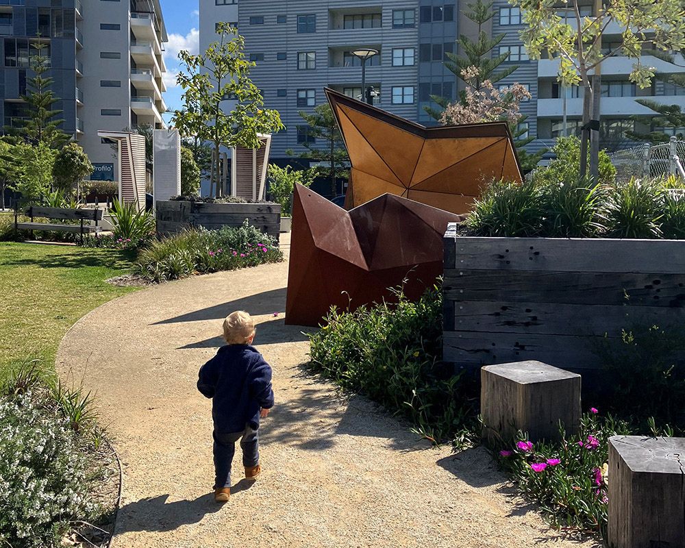 A photograph of a toddler walking on a crushed stone path, past trees in recycled timber planters. It's a sunny day. 