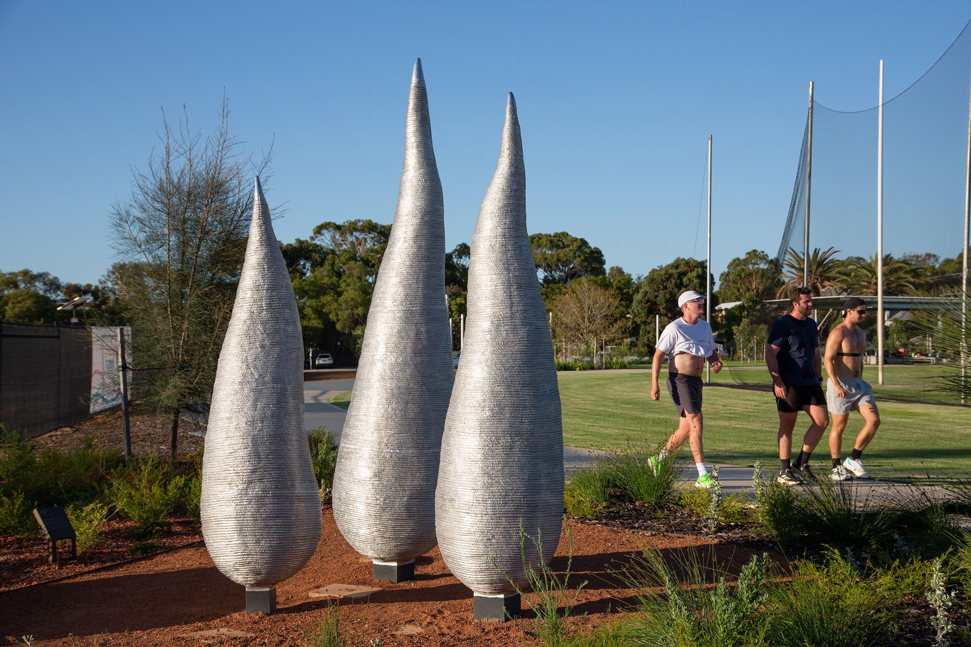 A photo of three organic, silver, sculptural forms in a park setting. Three people are walking in exercise gear on a path behind.