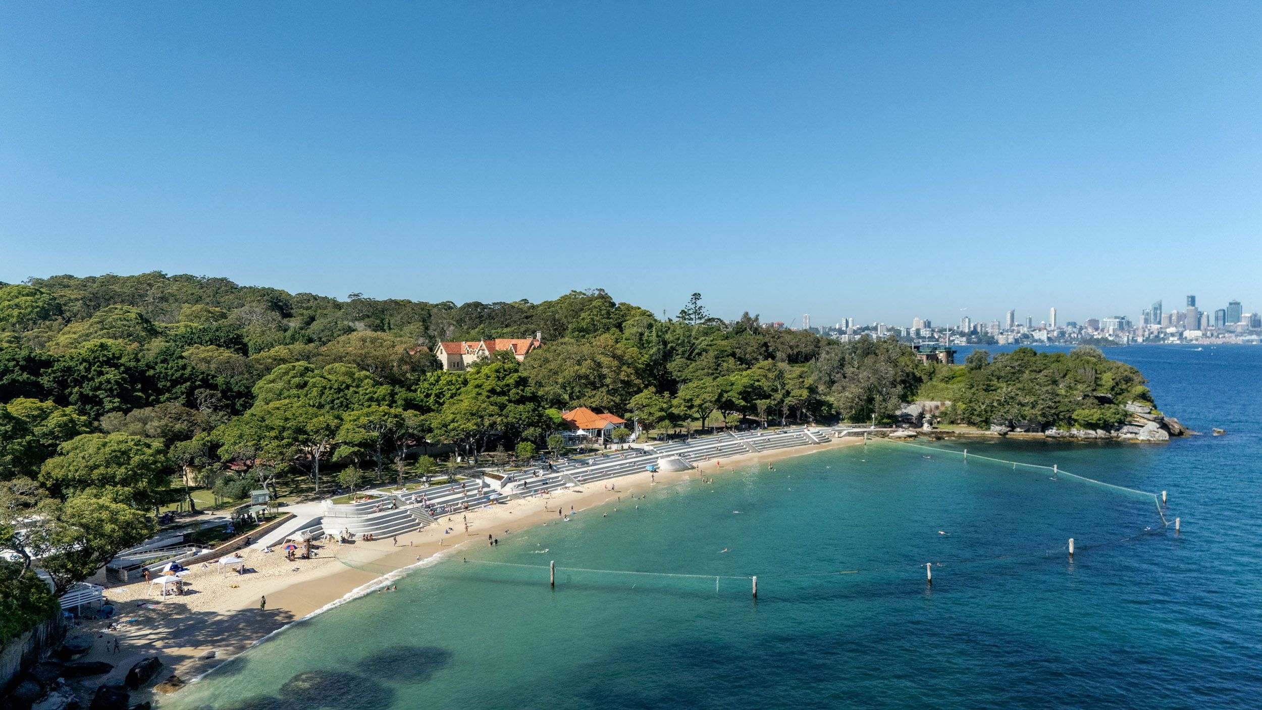 A drone photograph of a harbour beach featuring a tiered seawall and a shark net. The sky is blue and the beach is busy. A city skyline is in the distance.