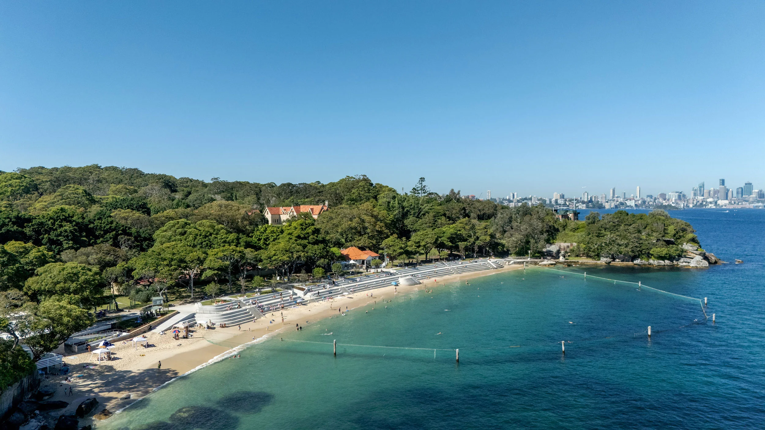 A drone photograph of a harbour beach featuring a tiered seawall and a shark net. The sky is blue and the beach is busy. A city skyline is in the distance.