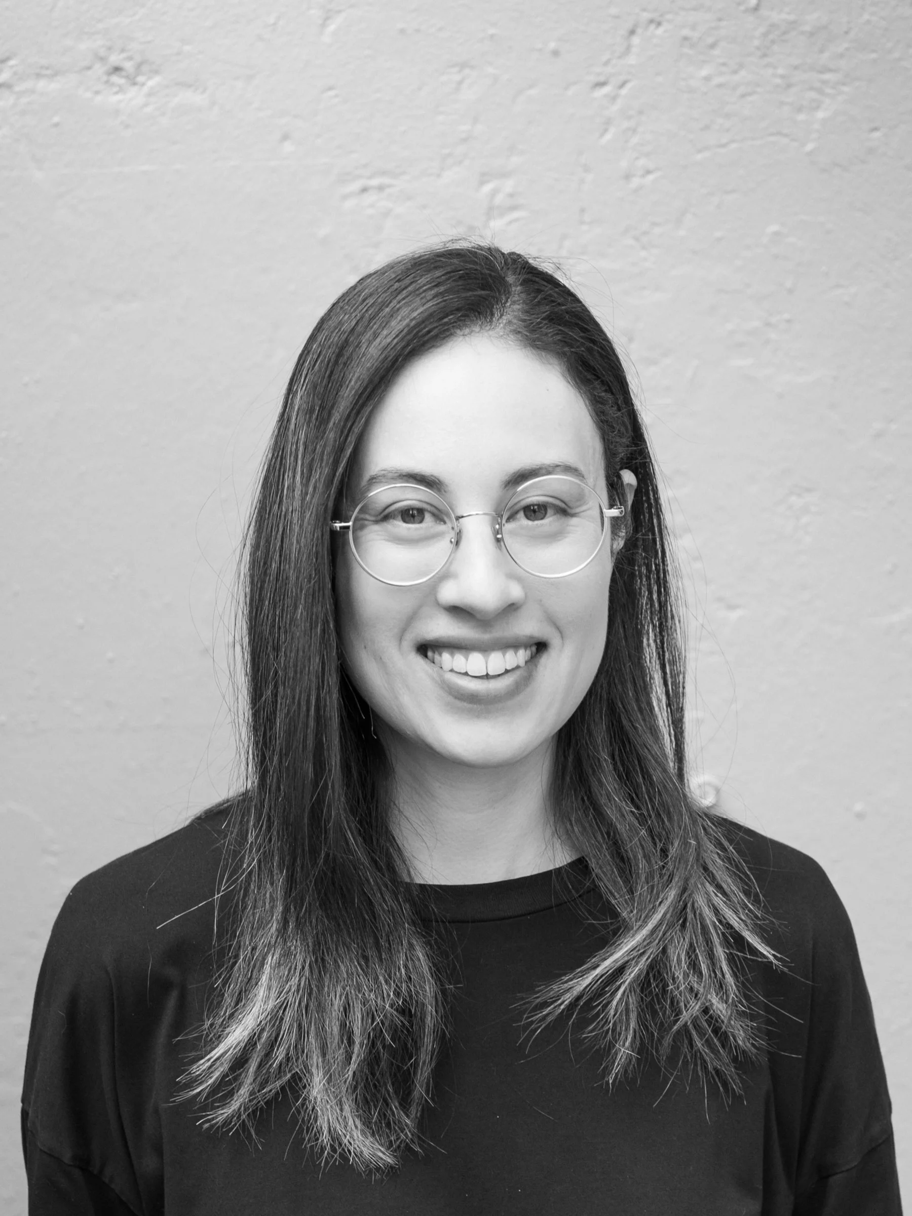 A black and white headshot of a person with long hair, glasses, and a big smile.