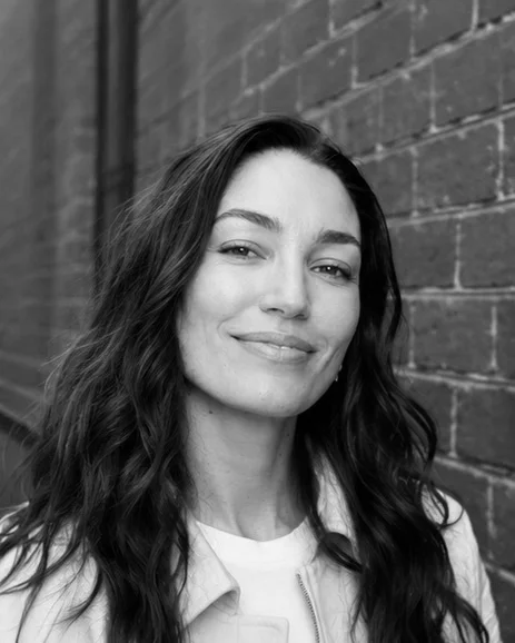 A black and white headshot of a person wearing a black top, standing in front of a brick wall.