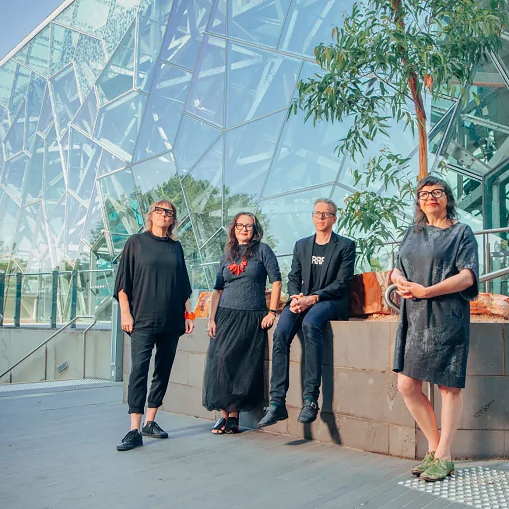 A photo of four people in front of a tree and a contemporary glass building.