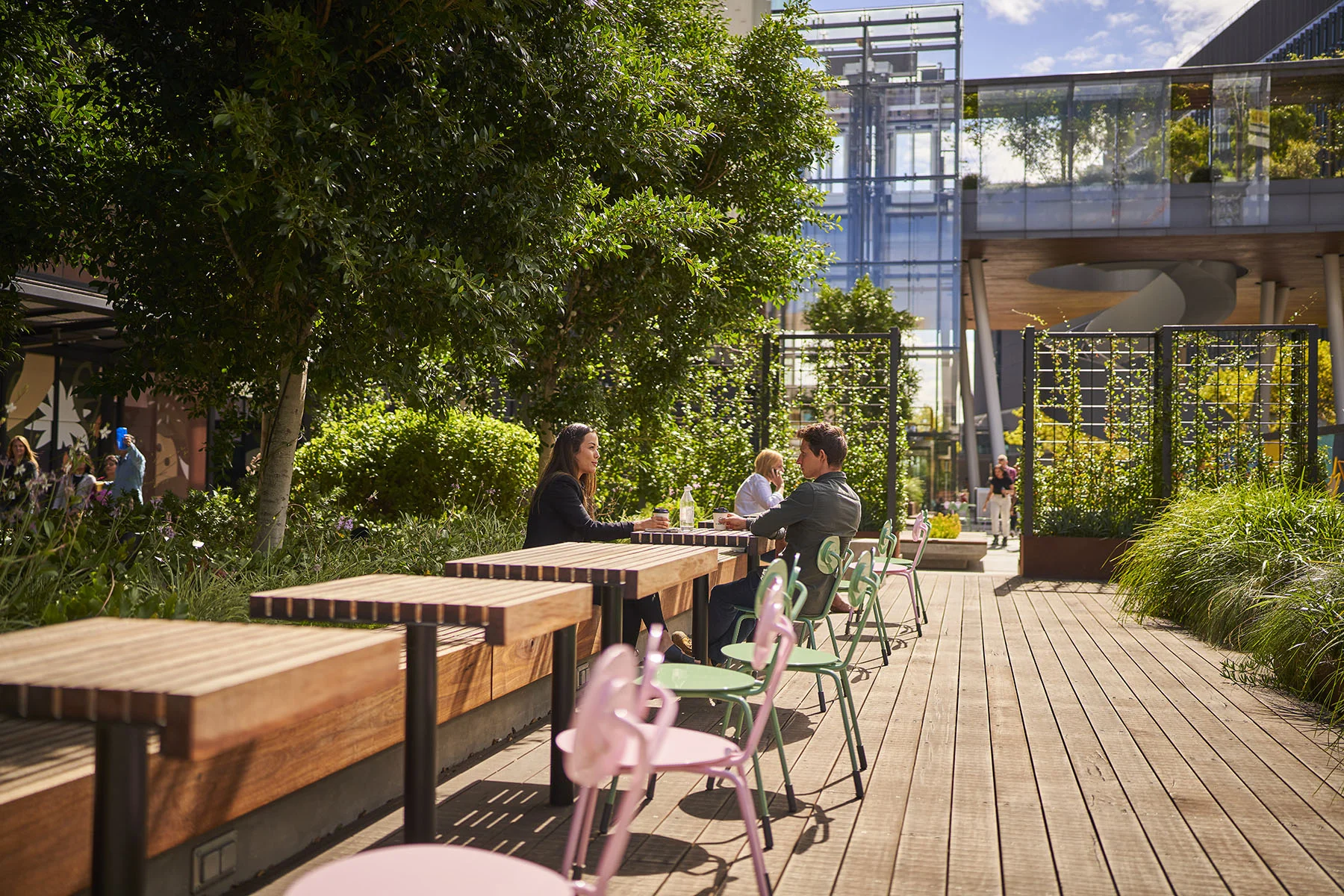 A photo of people at an outdoor seating area with timber benches plus pink and green chairs, surrounded by dense planting - trees, shrubs, and climbing vines.