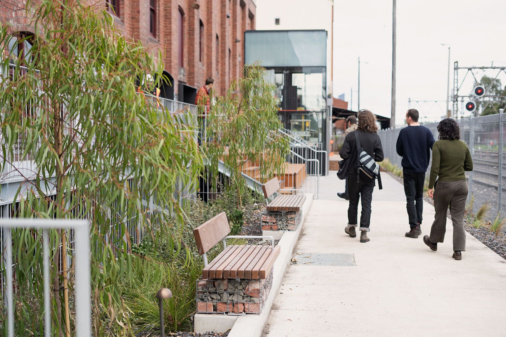 A photo of people walking down a path. On the right is a train line, on the left are trees and timber benches in front of an industrial building