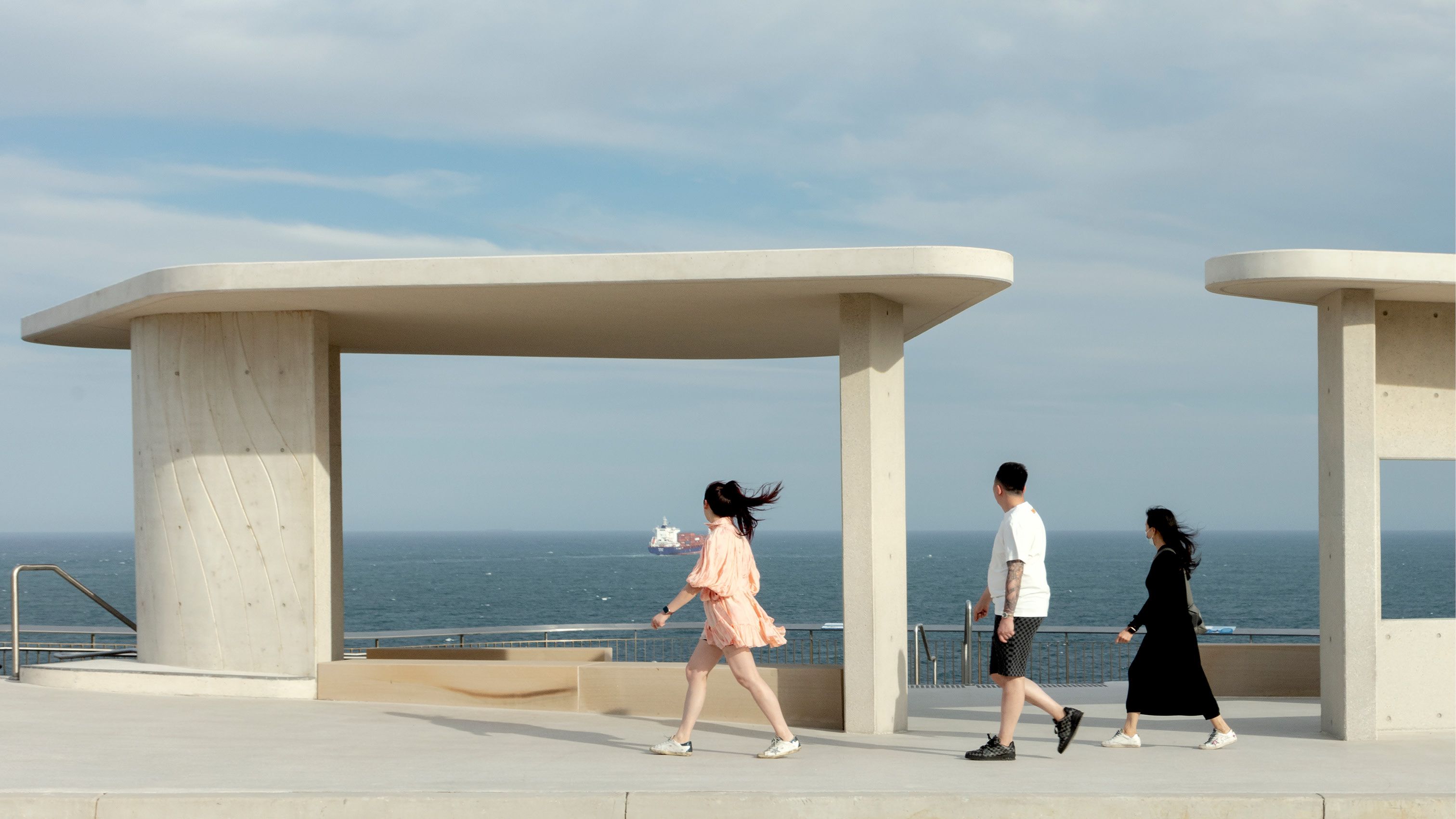 A photograph of three people walking in the wind in front of a lookout, looking out to sea. A container ship is visible in the distance. 