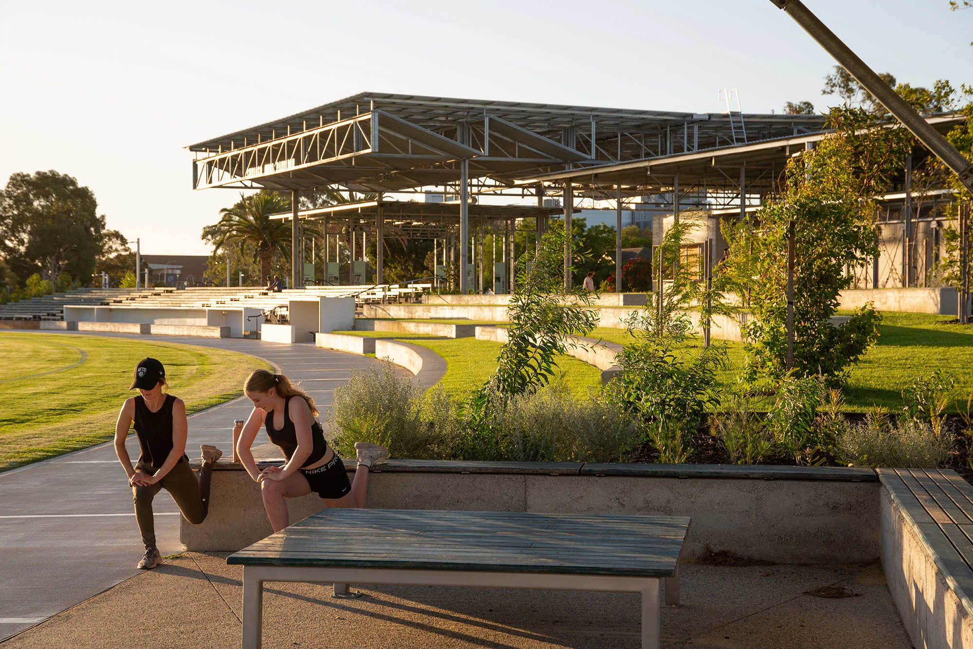 A photograph of two people stretching on a bench beside an oval. There is a pavilion with seating in the background.