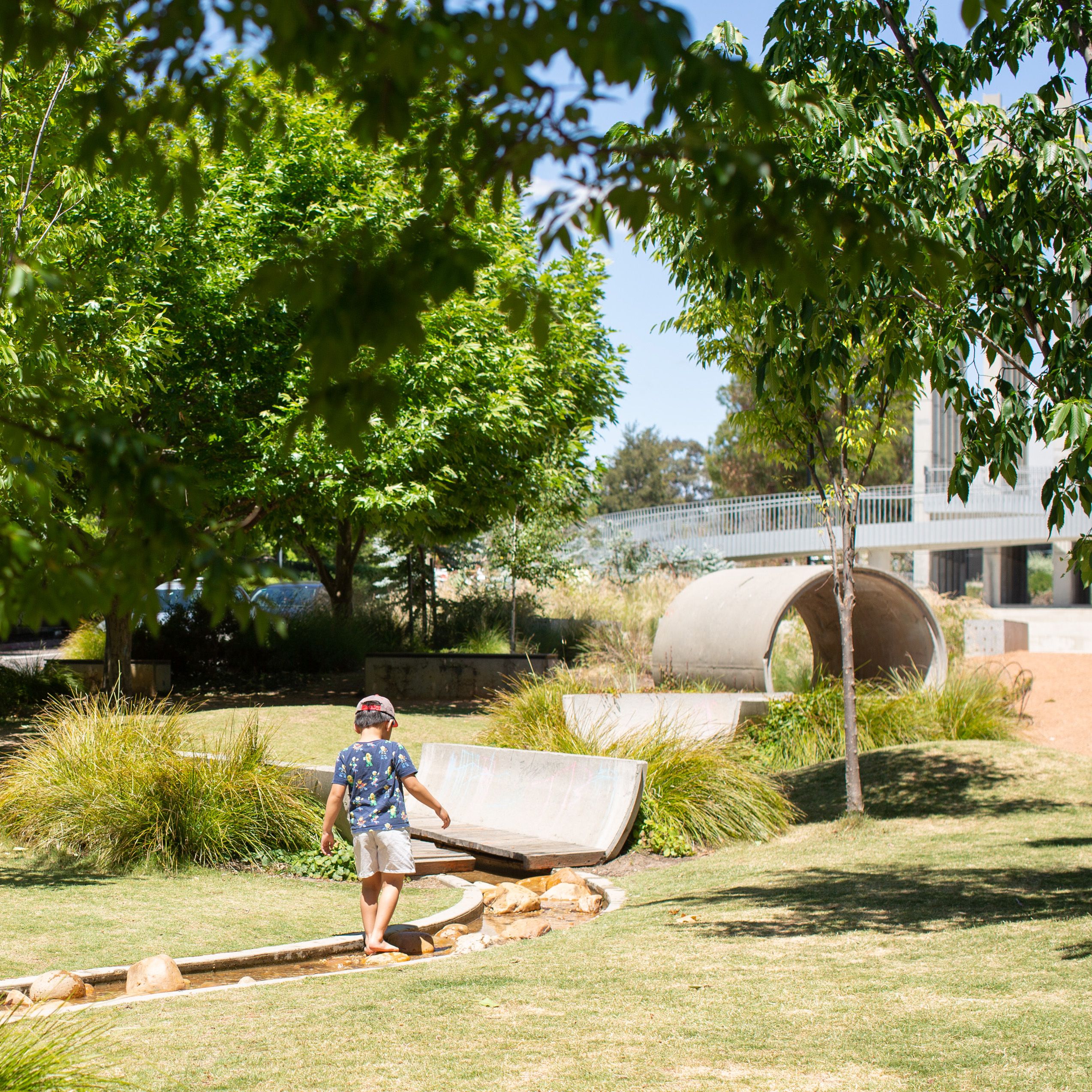A square photograph of an urban linear park landscape. There is a small child walking in the nature-play 'creek'. Industrial concrete forms are seen in the distance, including a large pipe for play, and a lush tree canopy obscures the blue sky.