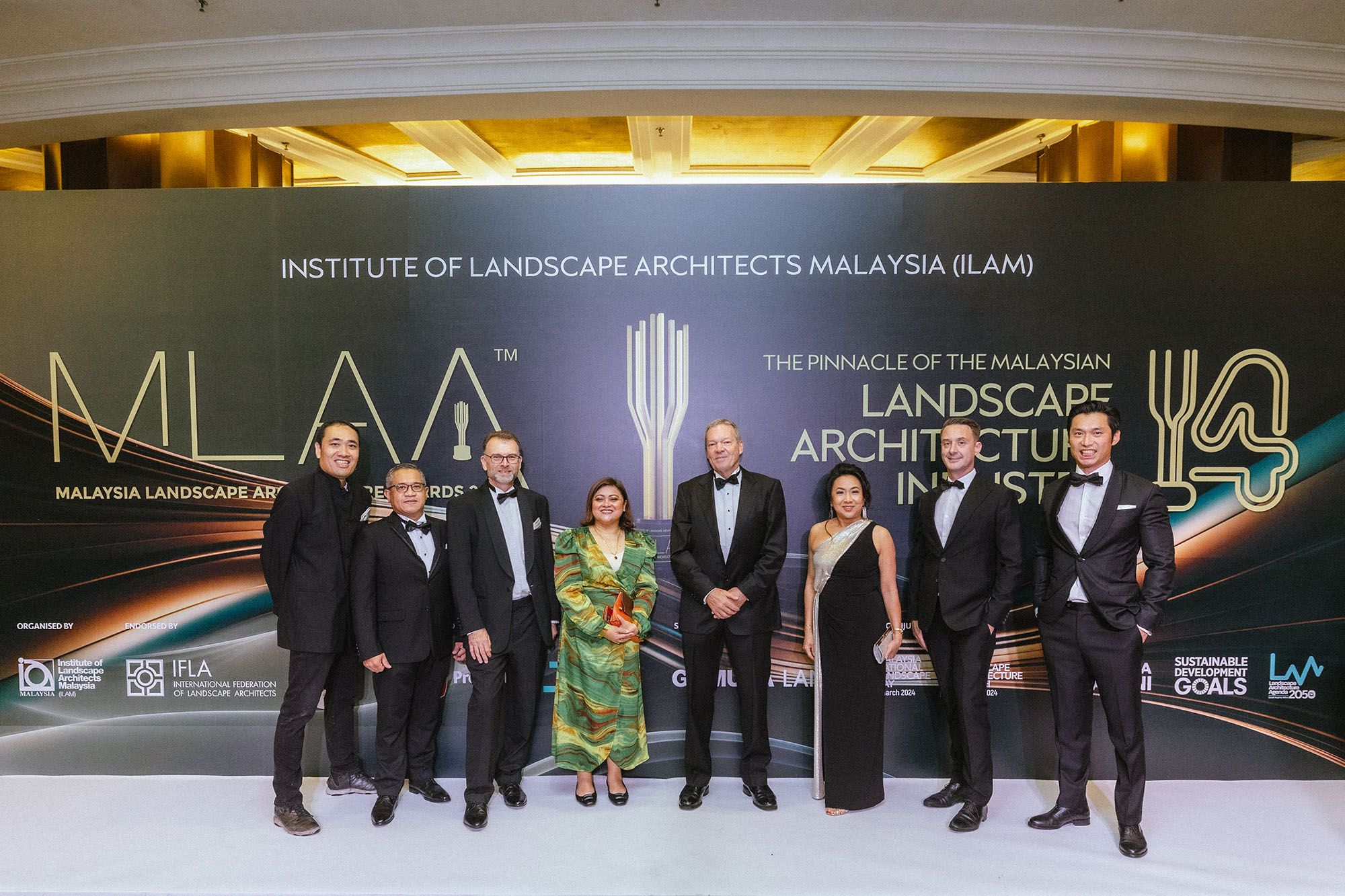 A photo of eight smiling people in formal attire, standing on a 'white carpet'