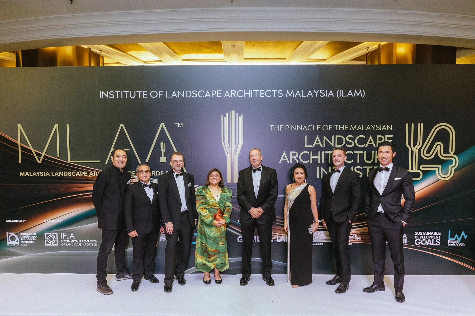 A photo of eight smiling people in formal attire, standing on a 'white carpet'