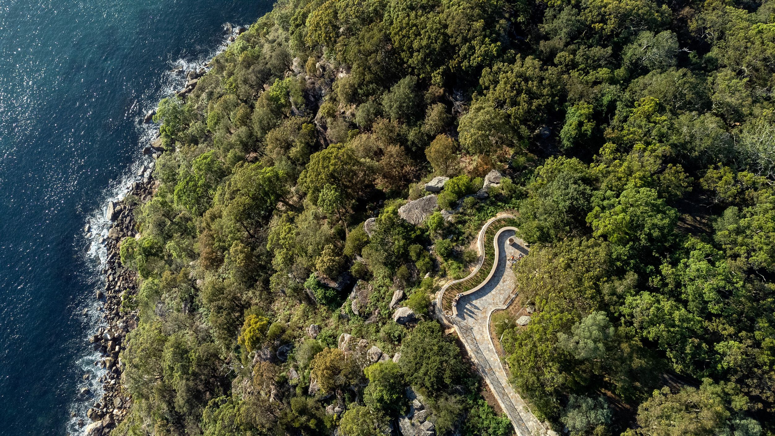 An aerial photograph of a curvy, sandstone lookout surrounded by green bushland, with water below.