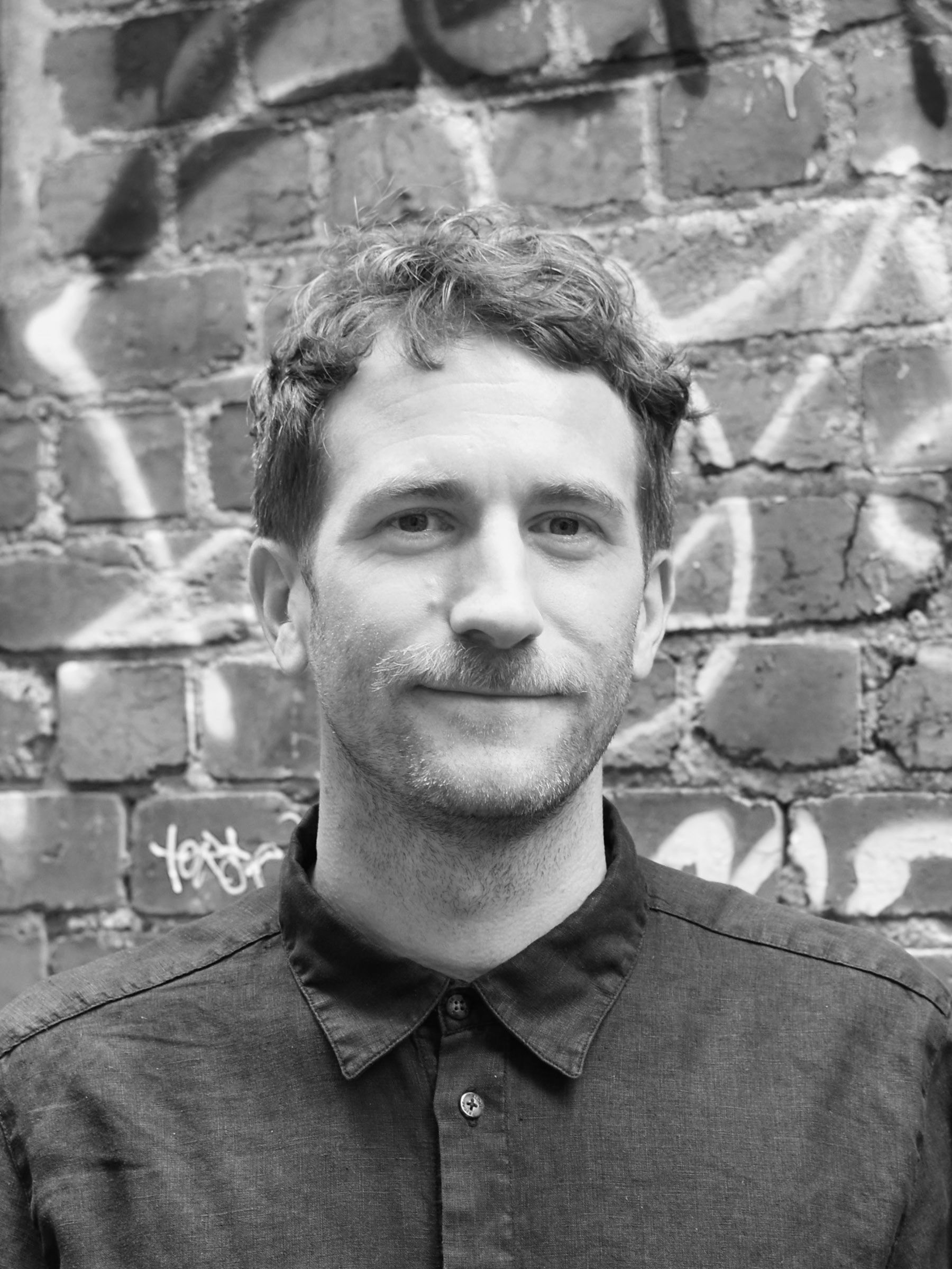 A black and white headshot of a person in front of a graffitied brick wall.