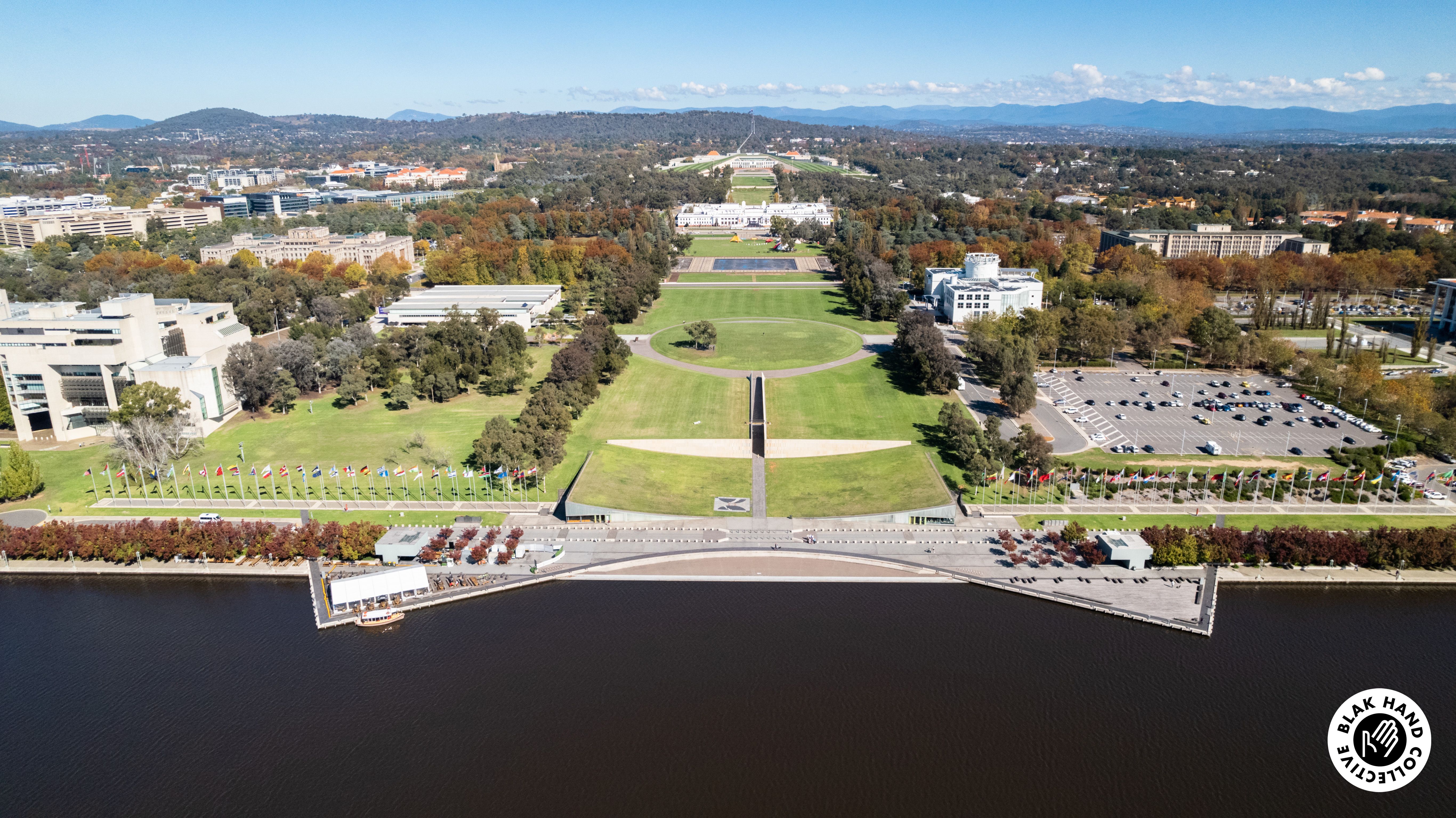 An aerial photograph of a lake front site. There is an large open corridor of grass running though the middle of the image, trees and buildings surround the open area, with mountains and blue sky visible at the top of the image. 