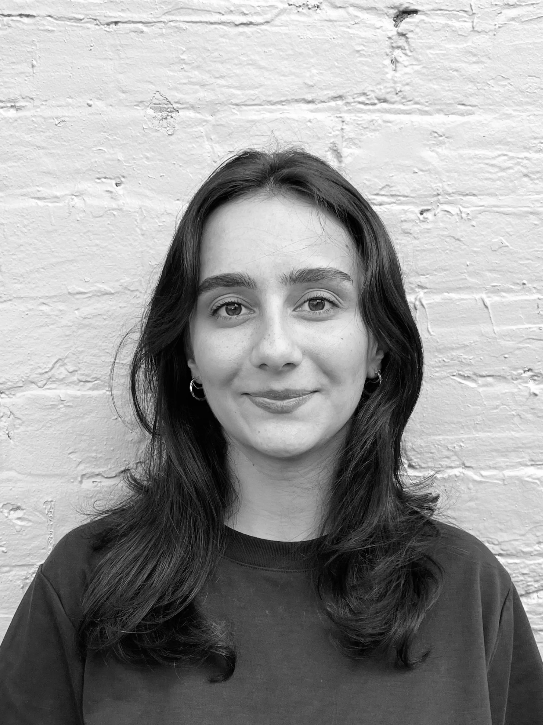 A headshot of a person with long hair standing in front of a brick wall. They are wearing a plain dark coloured t-shirt.
