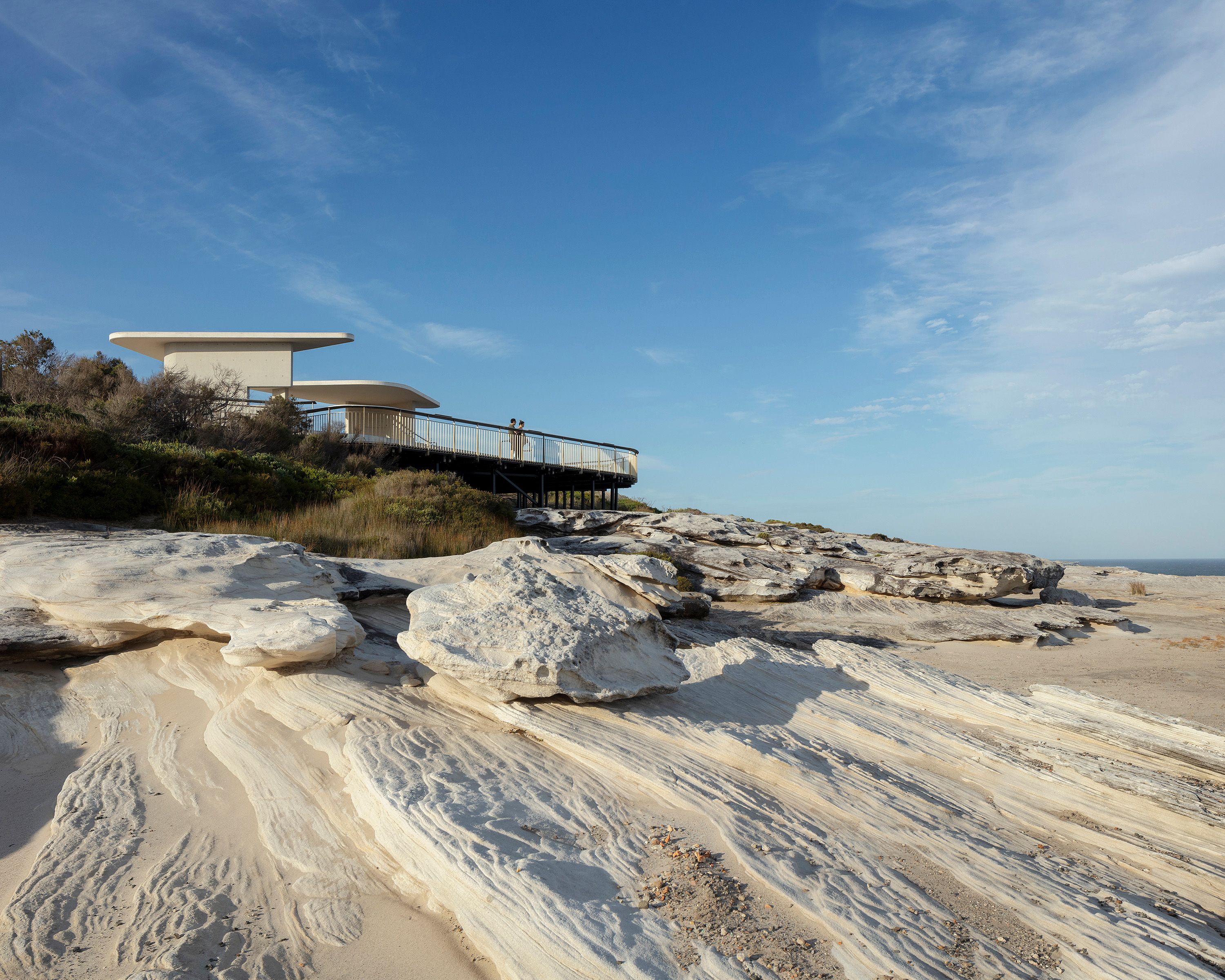 A photograph of a pale sandstone clifftop with a curved white concrete viewing shelter on top. The sky is bright blue with streaks of clouds. 