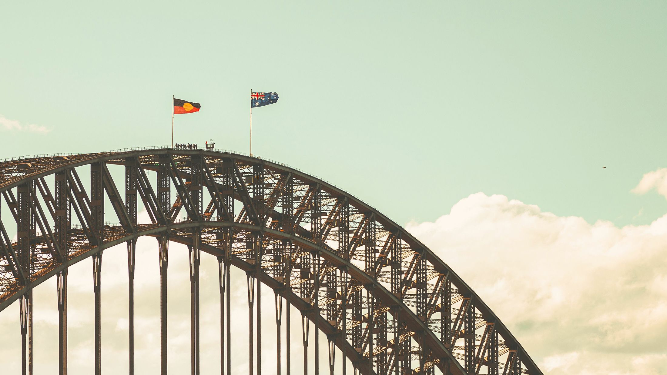 A photograph of the top of a steel arched bridge (The Sydney Harbour Bridge) with Aboriginal and Australian flags flying at full mast. There are a few people seen in silhouette near the top. 