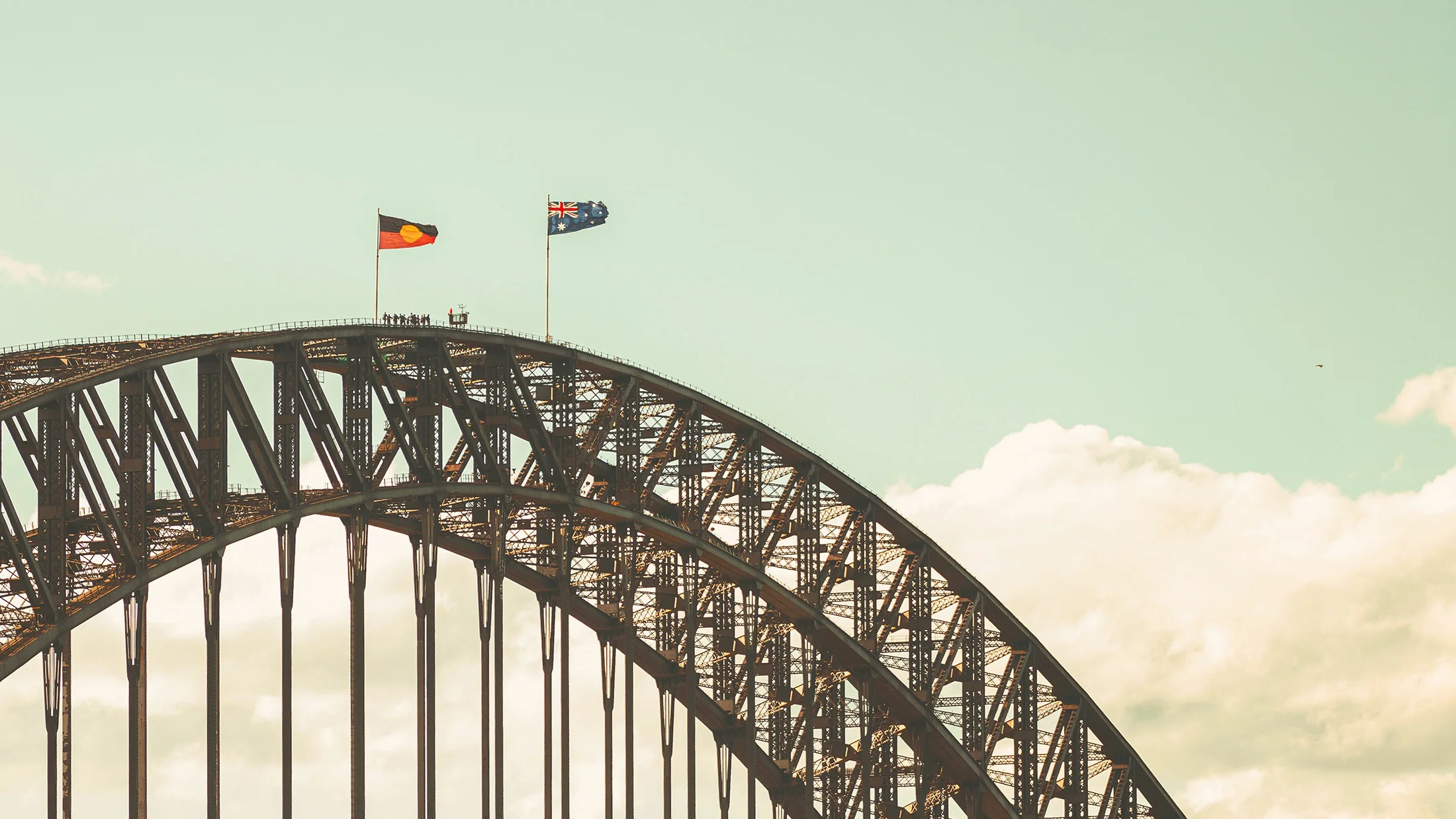 A photograph of the top of a steel arched bridge (The Sydney Harbour Bridge) with Aboriginal and Australian flags flying at full mast. There are a few people seen in silhouette near the top.