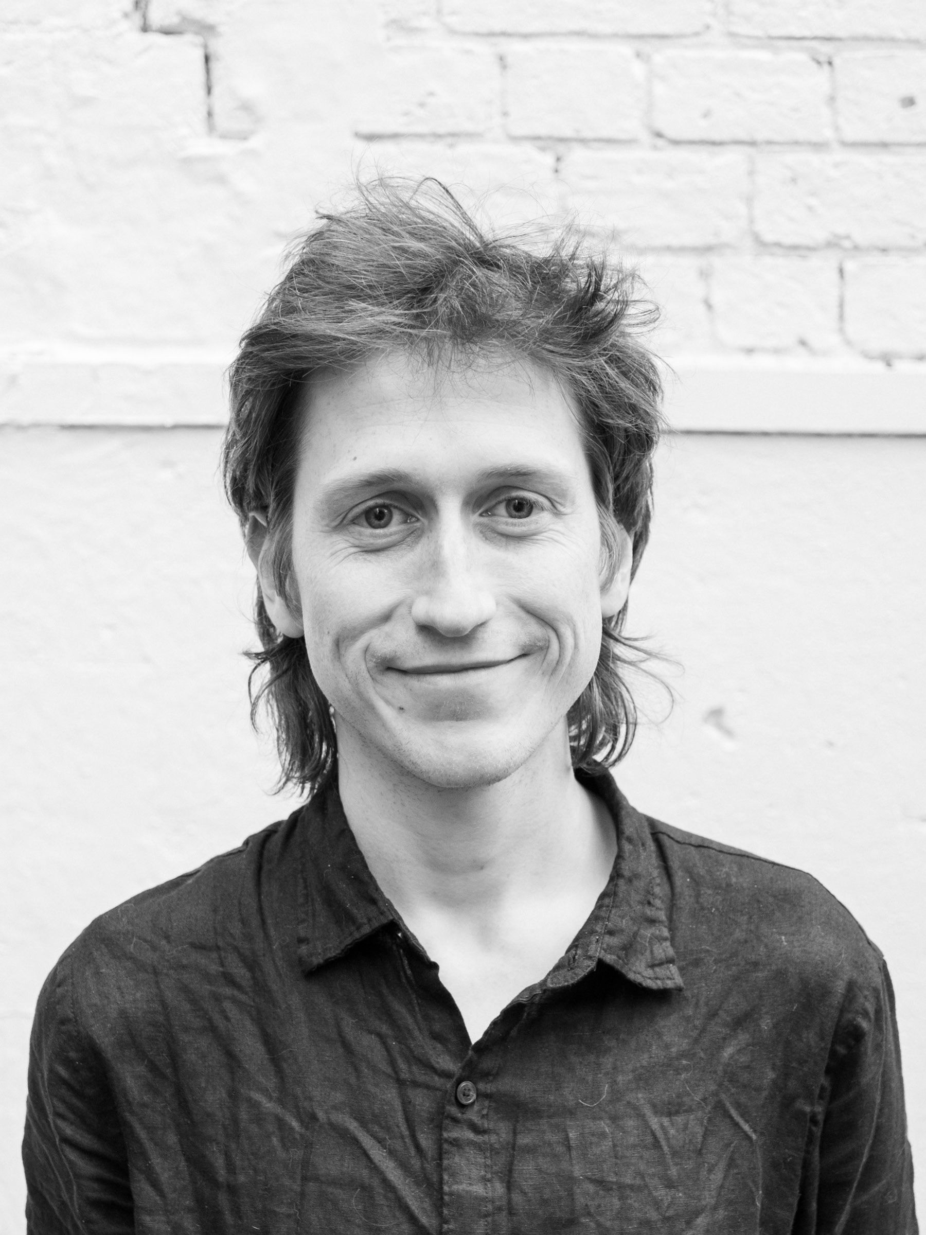 A headshot of a smiling person with short hair wearing a dark shirt, standing in front of a white wall.