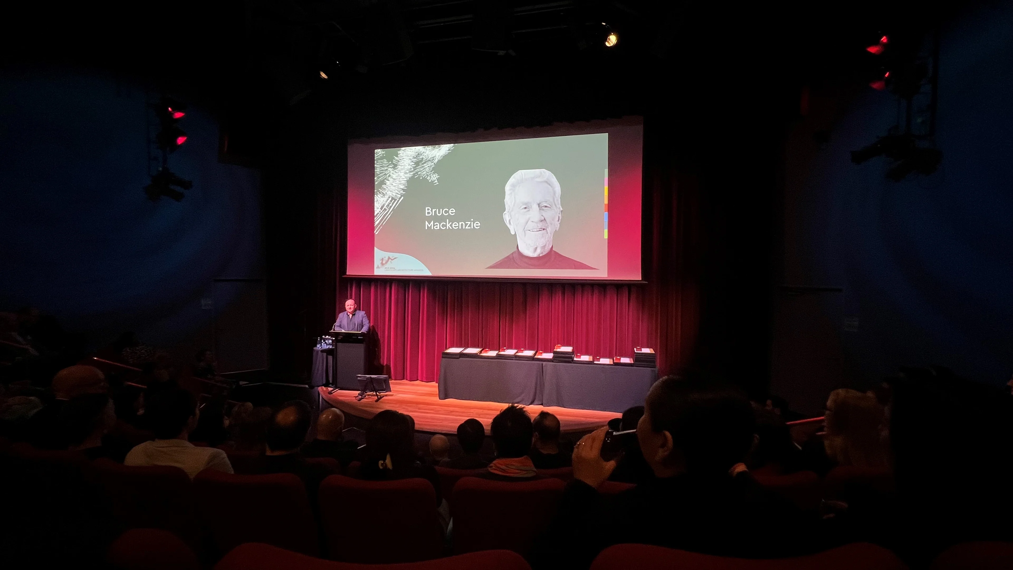 A photo of a stage, with one person at a lectern, and a black and white face projected above.
