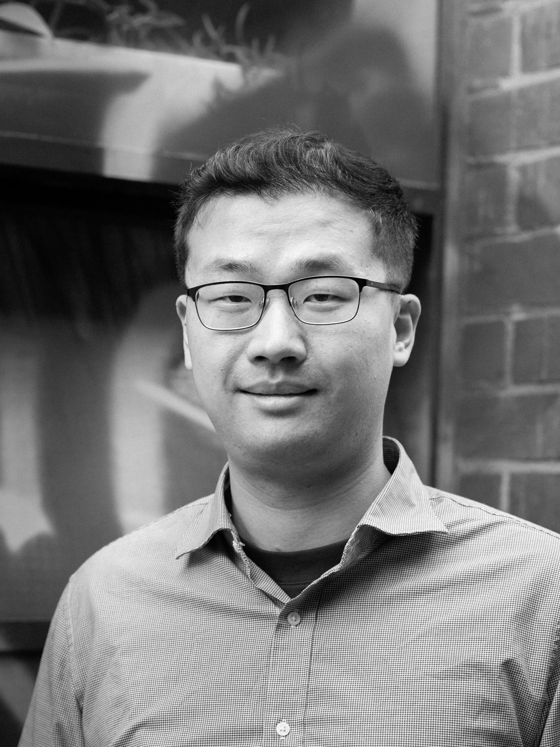 A black and white headshot of a person wearing glasses. The background is an gritty, urban wall.