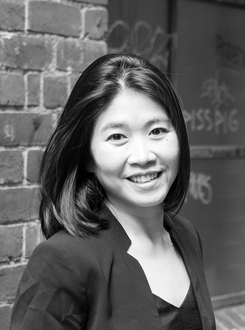 A black and white headshot of a smiling person with shoulder length hair in front of a brick wall.