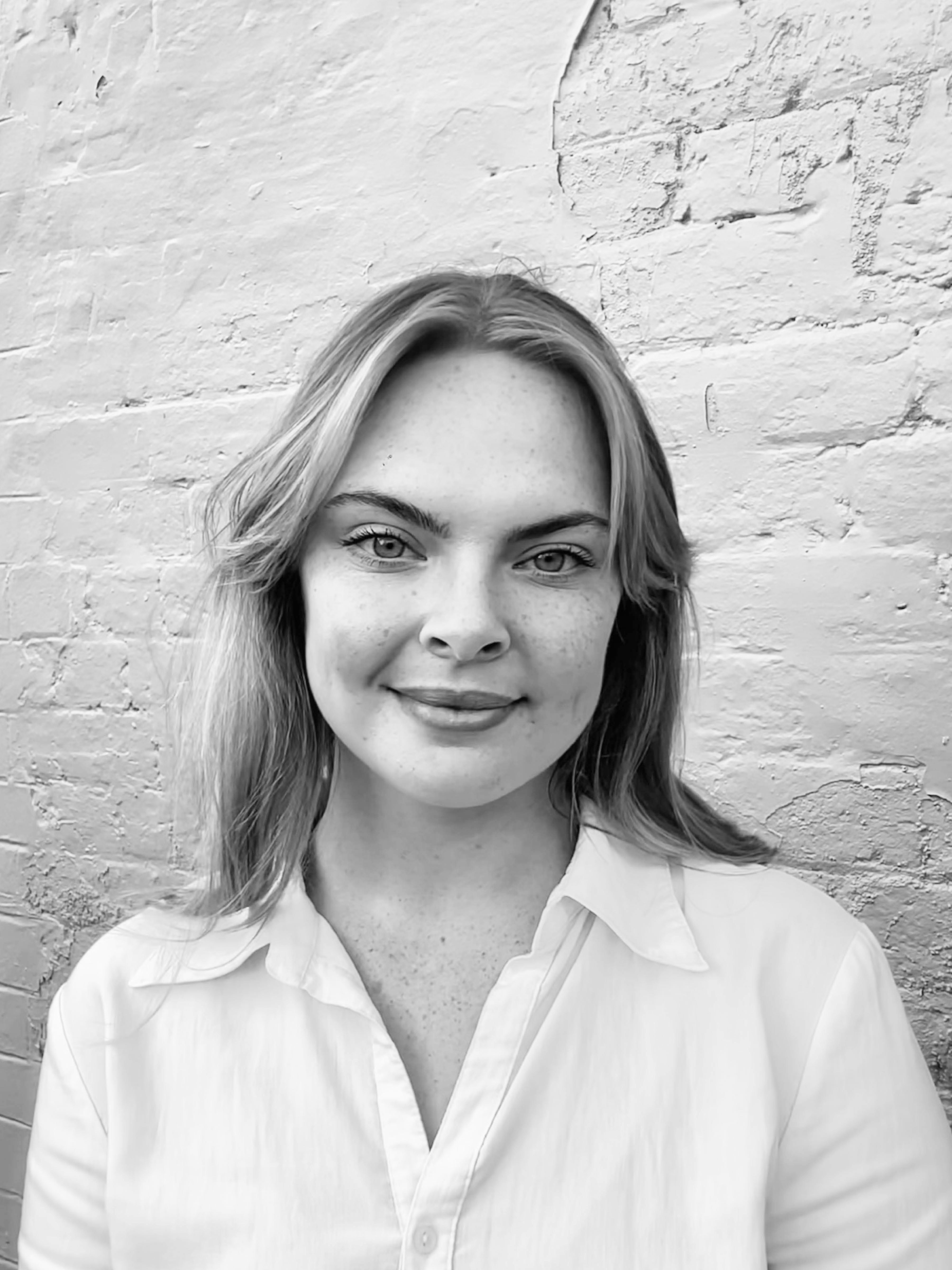 A black and white headshot of a person with long hair. They are wearing a white shirt and standing in front of a painted brick wall.