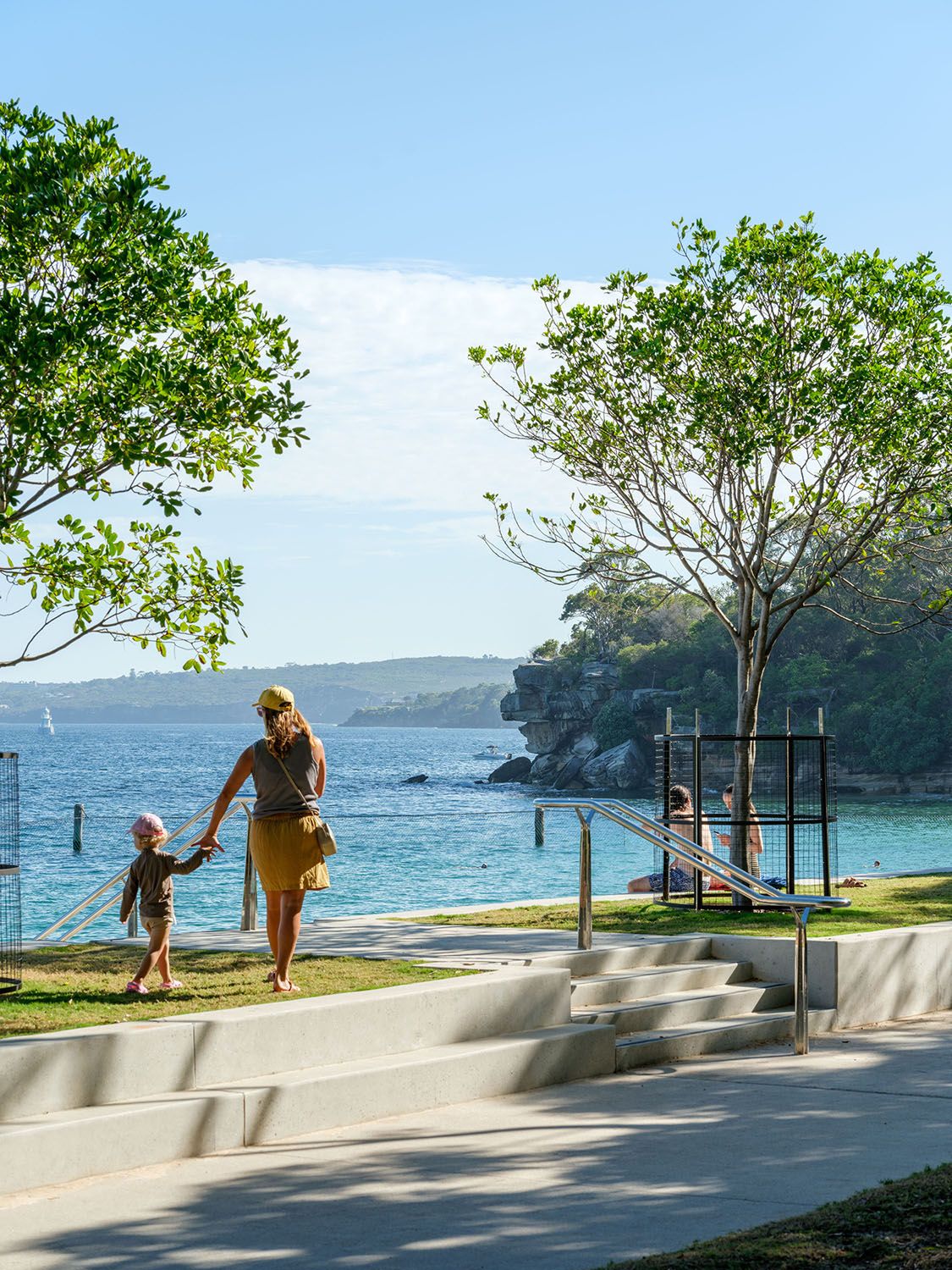 A photograph of an adult and child holding hands as they walk on a patch of lawn on top of a beachfront seawall. There is a tree either side, and other people obscured in the background. 
